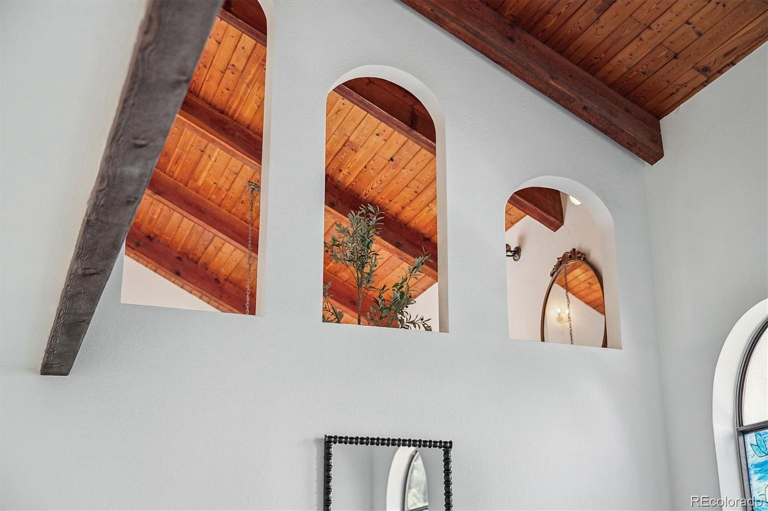 The image showcases a unique architectural detail with three arched niches in a white wall, offering glimpses of a wooden ceiling with beams. The niches are decorated with a plant and mirrors, adding depth and visual interest. The overall style is rustic and charming, highlighting the home's character.