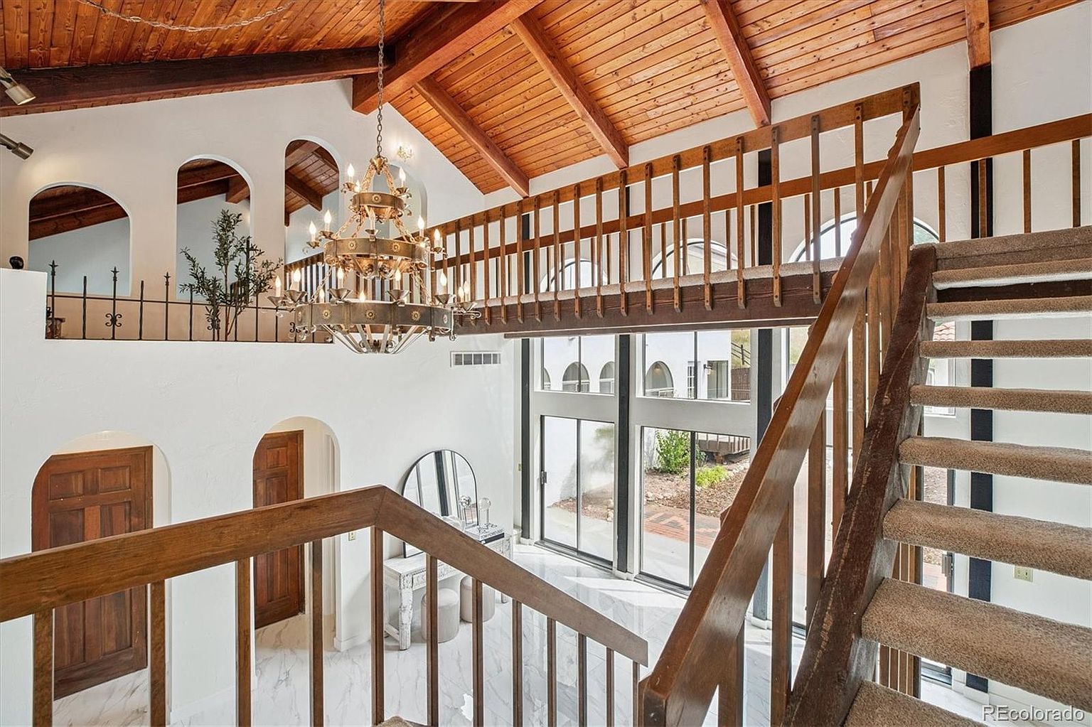 This interior shot showcases a grand hallway and staircase. The space features a high, wood-paneled ceiling, a large chandelier, and a balcony overlooking a lower level with large windows. The staircase is carpeted and has wooden railings, contributing to the home's elegant and spacious feel.