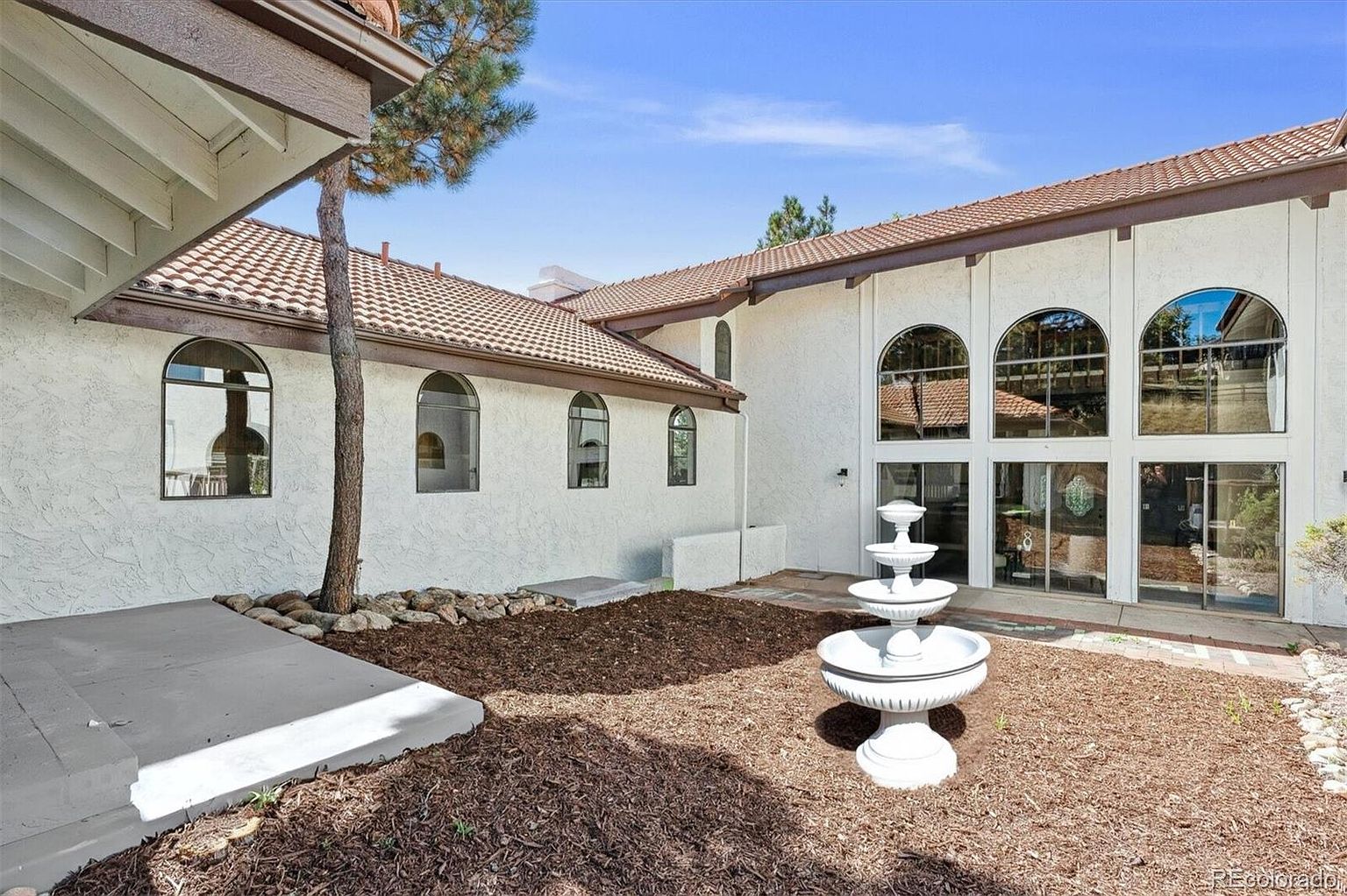 This is a rear exterior view of a two-story home featuring a white stucco facade and a terracotta tile roof. Large arched windows provide ample natural light, and a tiered fountain adds a touch of elegance to the landscaped yard. The overall impression is one of sophisticated charm and well-maintained property.