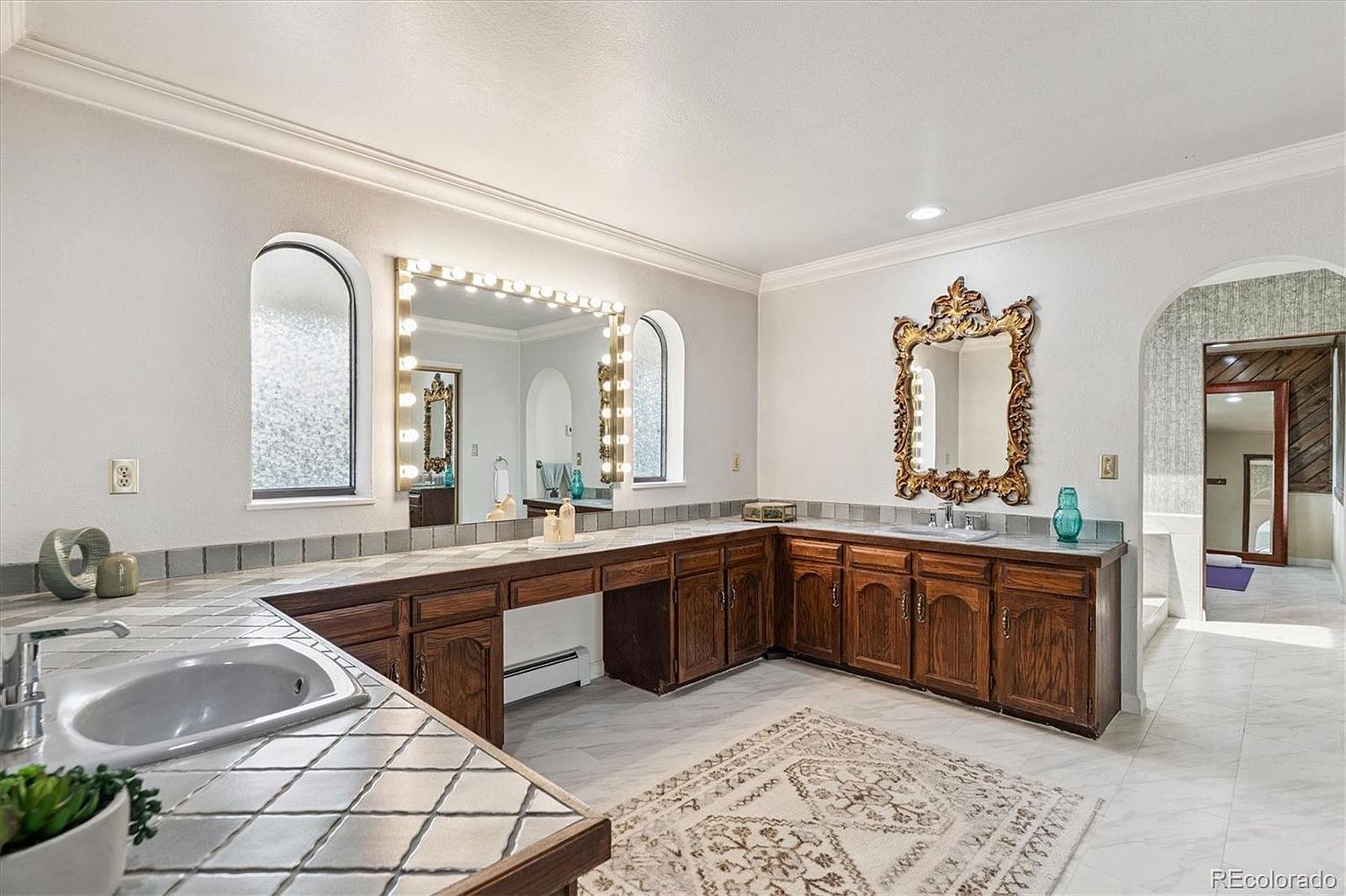 This is a spacious primary bathroom featuring a long vanity with dark wood cabinets, a tile countertop, and two sinks. A large mirror with Hollywood-style lighting is above the vanity, and an ornate mirror hangs above the second sink. The floor is tiled, and a decorative rug adds warmth to the space.