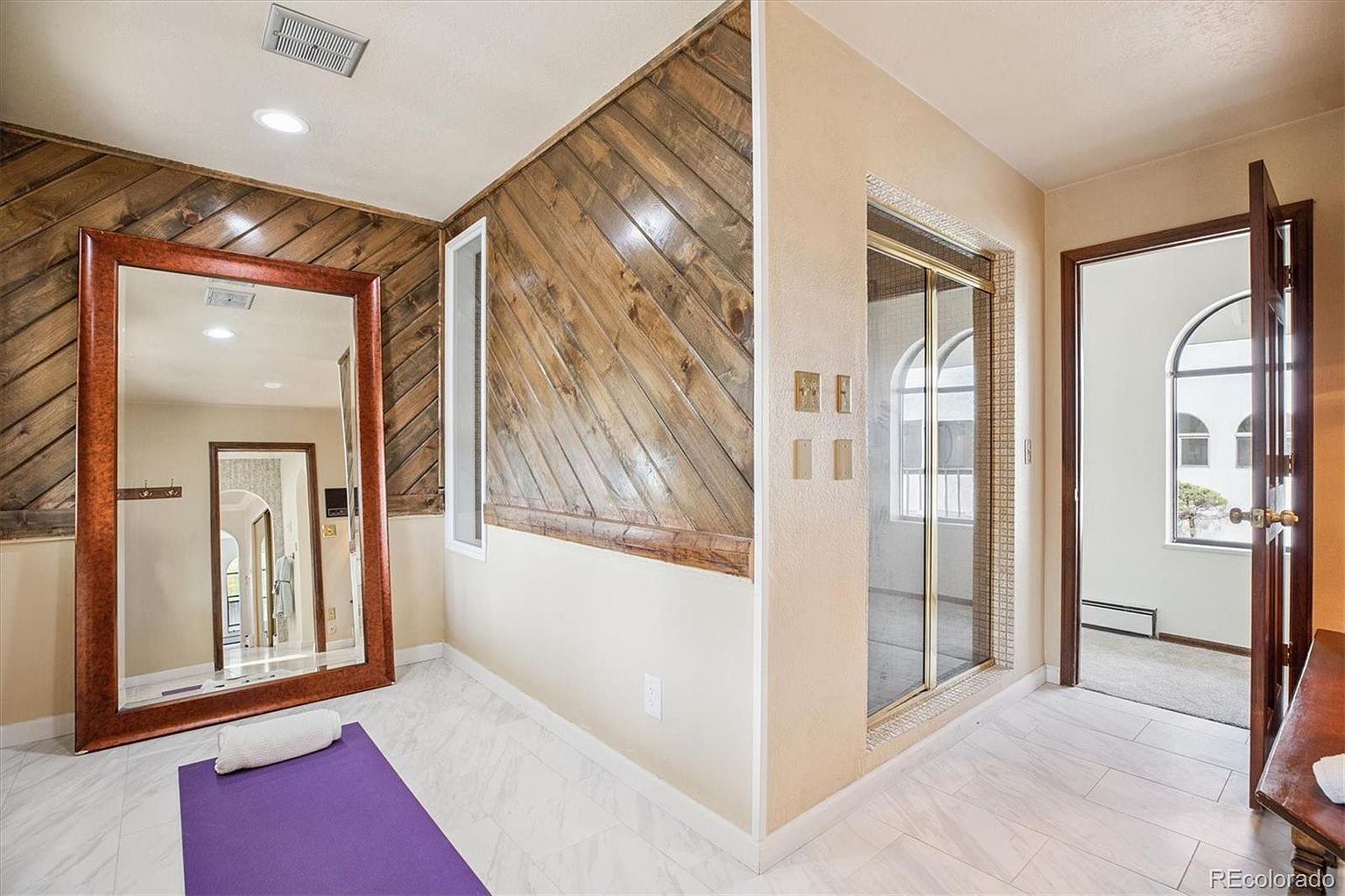 This interior shot showcases a home gym area with a large mirror, wood-paneled accent wall, and a purple yoga mat on a white tile floor. A shower enclosure is visible in the background, along with a doorway leading to another room, suggesting this space is part of a larger bathroom or wellness area. The style is a blend of rustic and modern, creating a unique and inviting workout space.