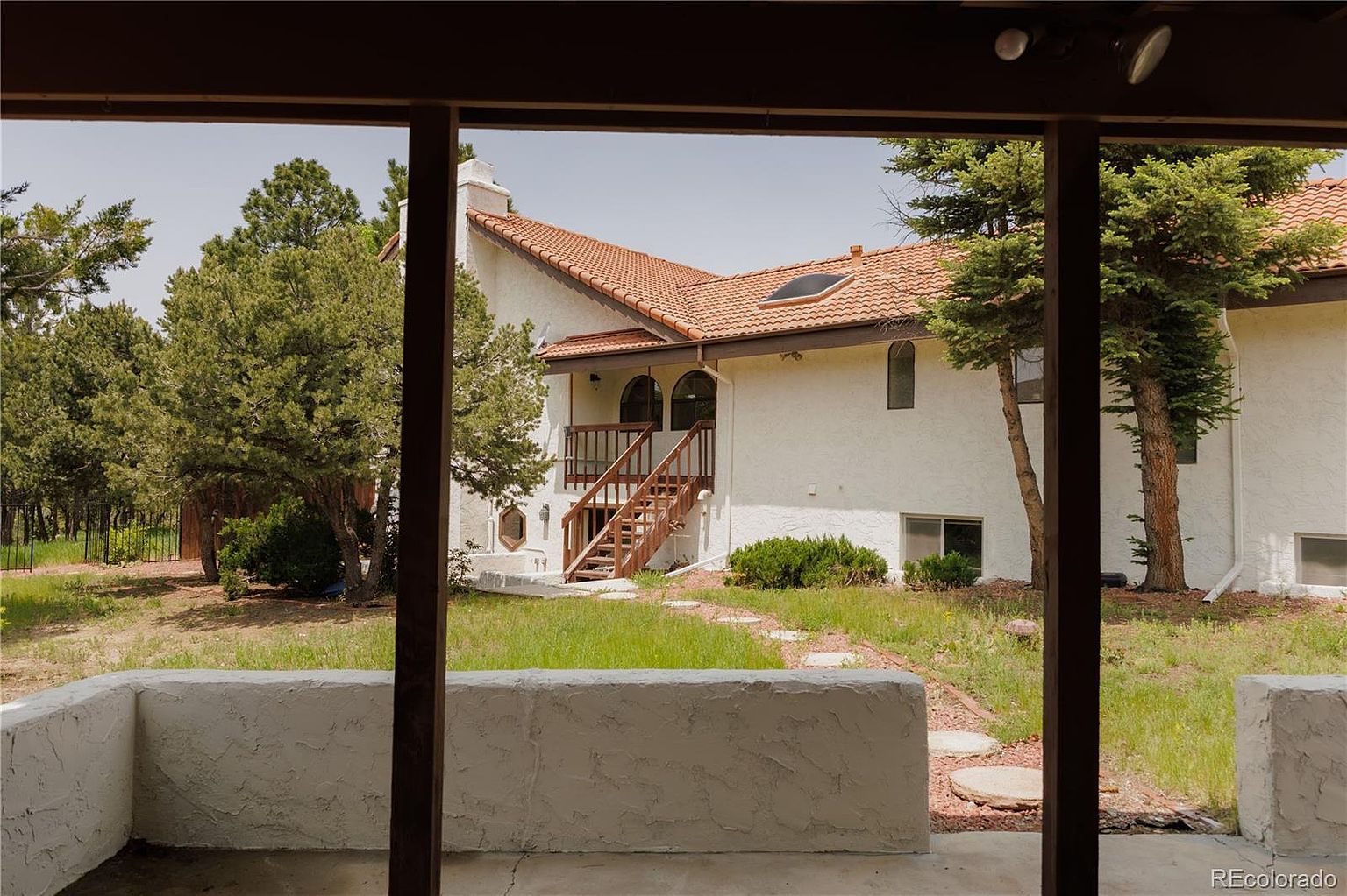 This image showcases the rear exterior of a two-story house with a terracotta tile roof and white stucco walls. A wooden staircase leads to a balcony, and mature trees surround the property, adding to its privacy and charm. The view is framed by a covered patio area, offering a glimpse of the outdoor living space.