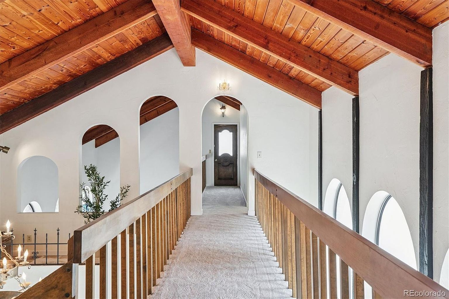 This interior shot showcases a hallway with a distinctive architectural design, featuring arched doorways and a warm, wooden ceiling with exposed beams. A wooden railing lines the hallway, leading towards a dark wood door at the end, while the walls are painted in a neutral tone, creating a bright and inviting space. The carpeted floor adds a touch of comfort and elegance.