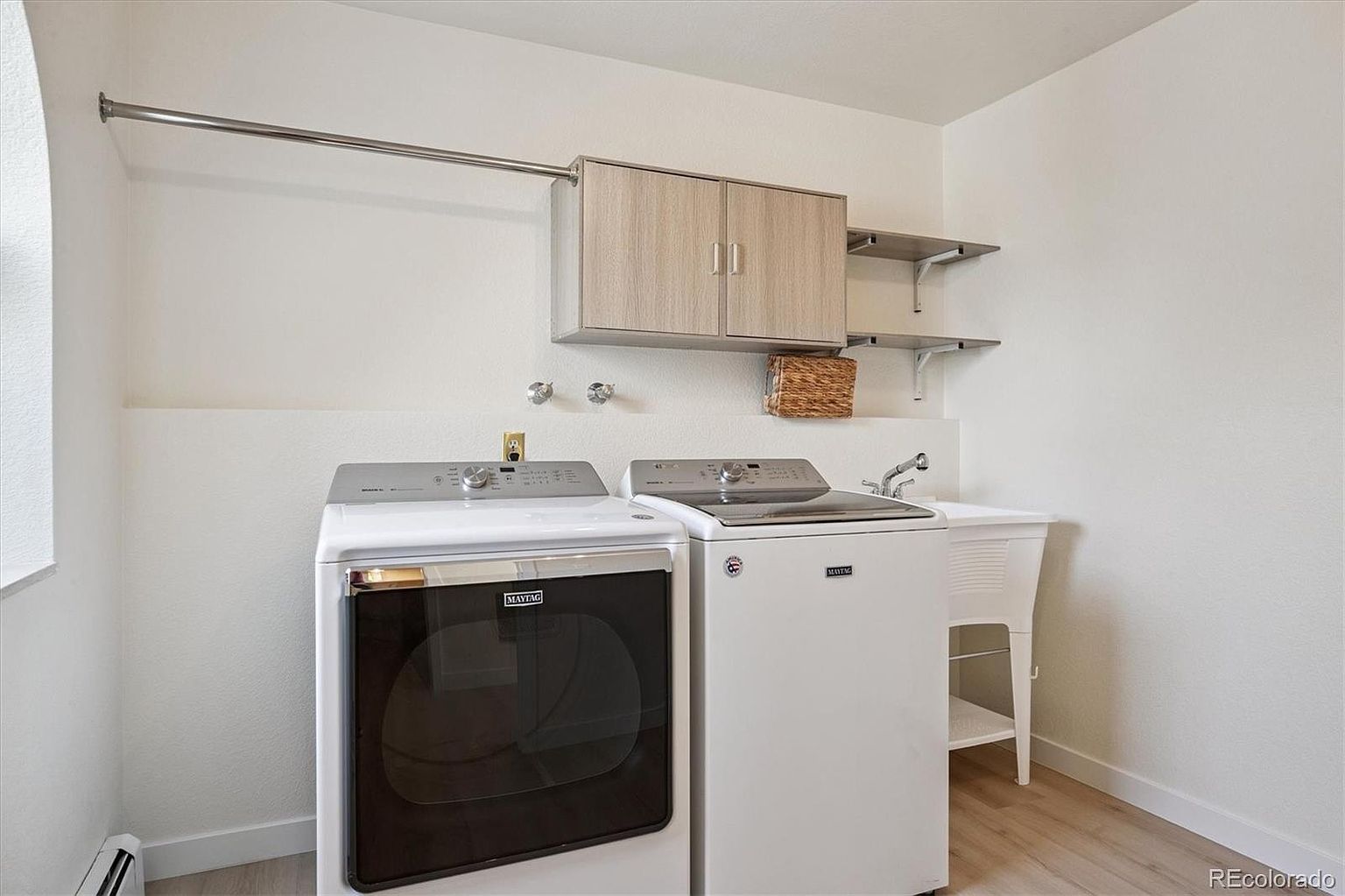 This is a well-organized laundry room featuring a front-load washer and top-load dryer, both in white. Above the appliances, there's a cabinet for storage and a hanging rod for clothes. A utility sink with a faucet is located to the right of the dryer, and the room has light wood-look flooring, creating a clean and functional space.