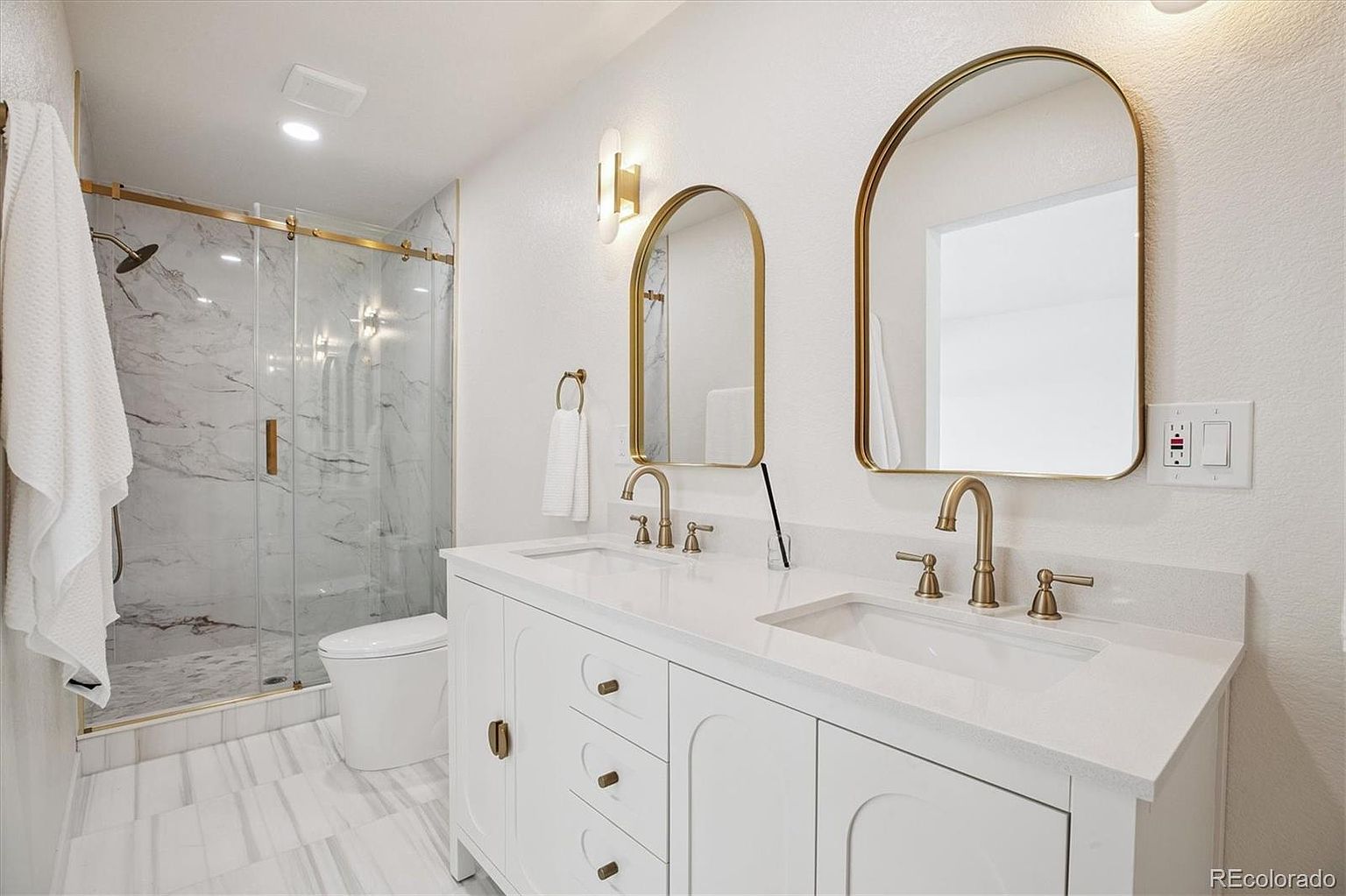 This is a bright and modern primary bathroom featuring a double vanity with white cabinetry and gold hardware. The countertops are a clean white quartz, complemented by gold-framed arched mirrors and matching fixtures. A glass-enclosed shower with marble-like tiling adds a touch of luxury, and the overall aesthetic is clean, elegant, and well-lit.