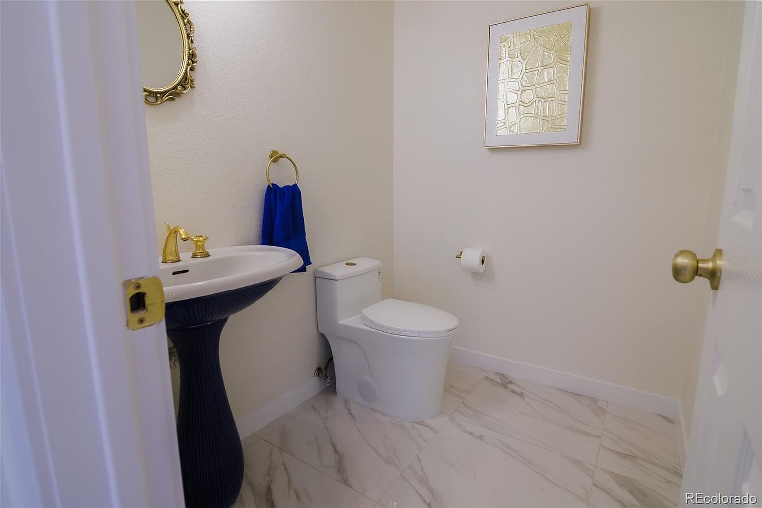This is a well-lit guest bathroom featuring a pedestal sink with gold fixtures, a white toilet, and marble-patterned tile flooring. A gold-framed mirror and artwork add a touch of elegance to the space. The room appears clean and modern, with a focus on classic design elements.