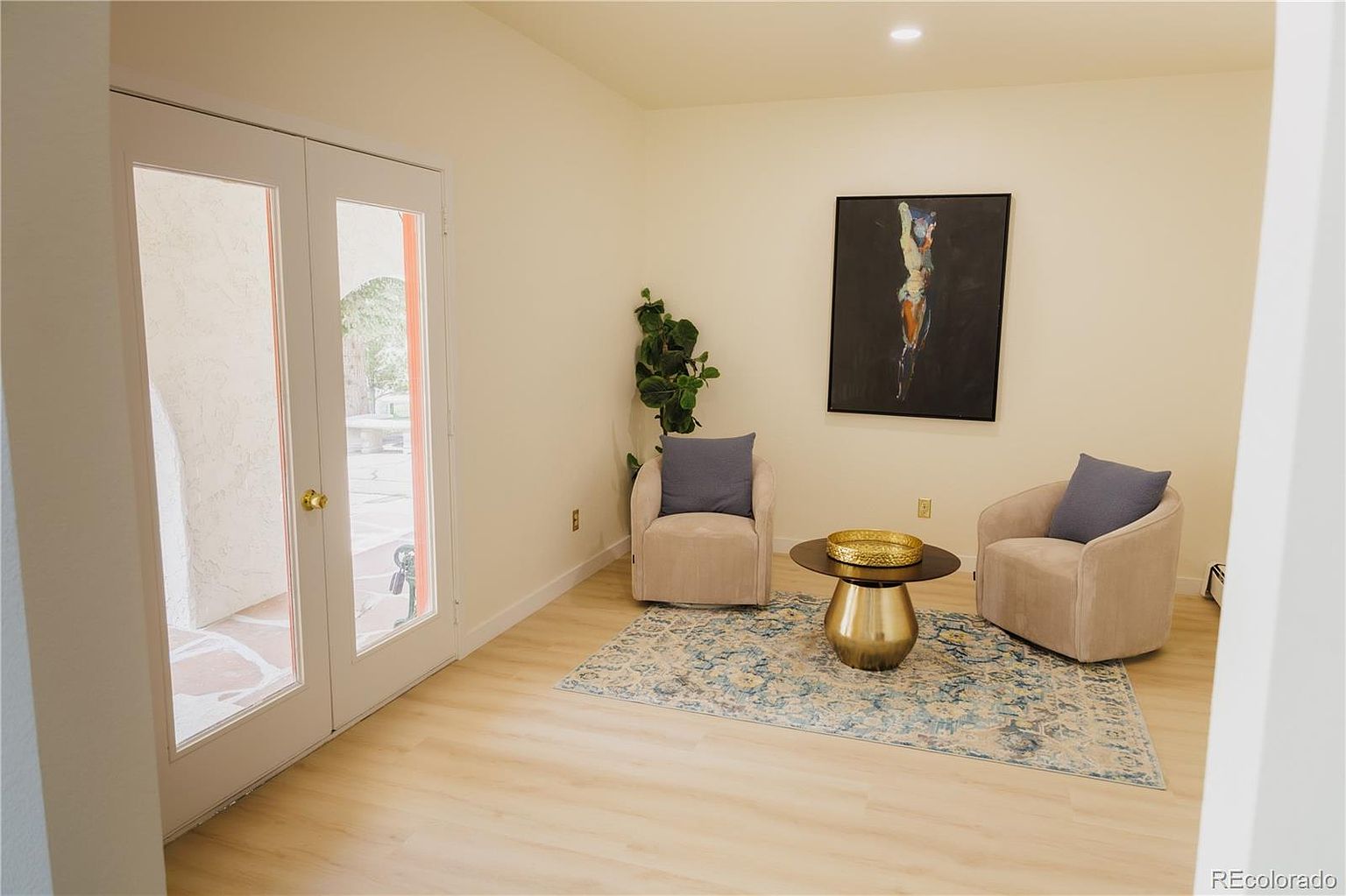 This is an interior shot of a cozy living room featuring two armchairs, a round coffee table, and a piece of art on the wall. The room has light-colored walls and flooring, creating a bright and airy atmosphere. French doors lead to an outdoor area, enhancing the sense of space and connection to the outside.