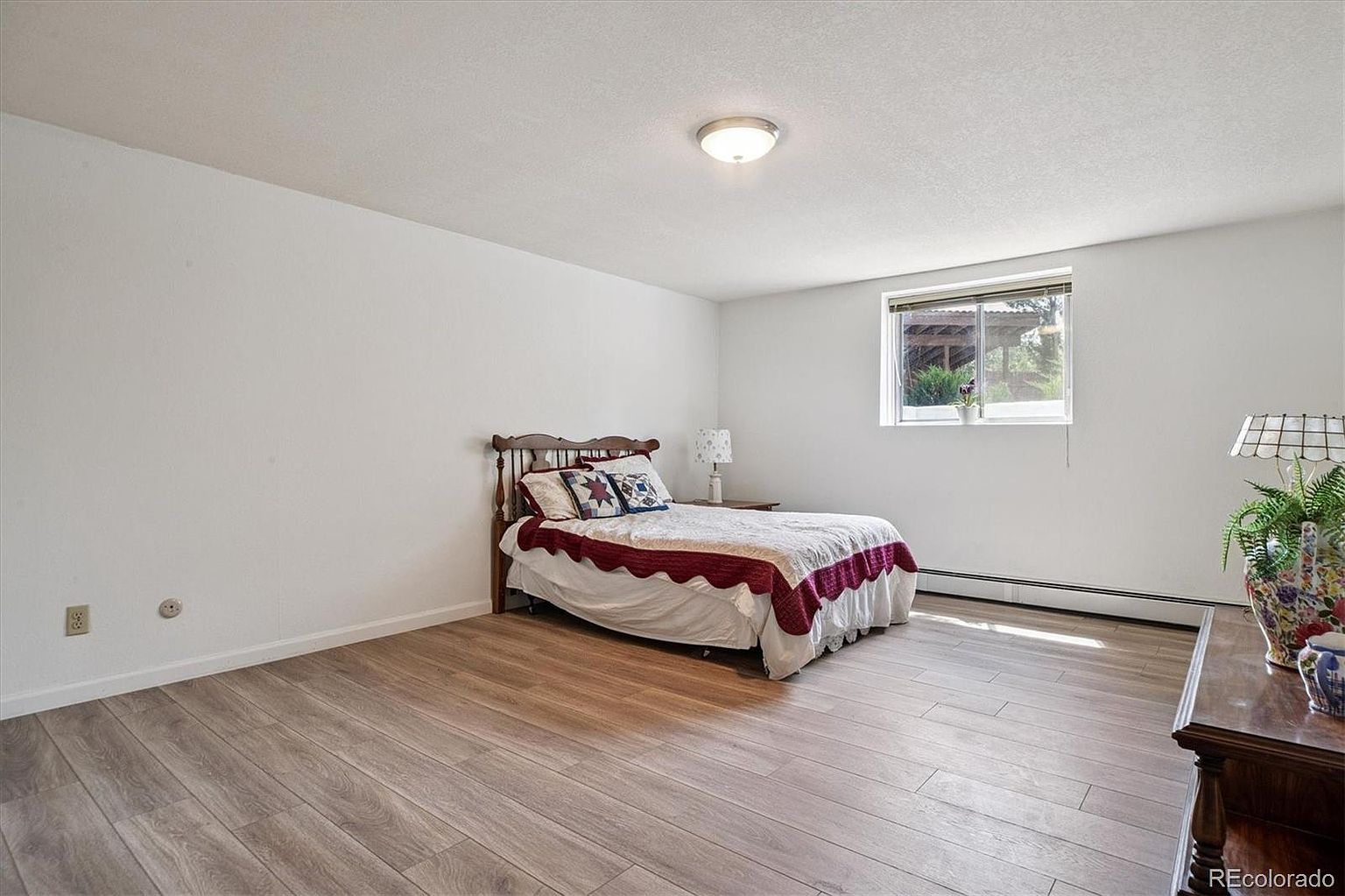 This is a bright and airy bedroom featuring light wood-look flooring and white walls. A bed with a wooden headboard and decorative pillows is centered in the room, complemented by a small side table and lamp. A window provides natural light, and a dark wood dresser is visible on the right side of the frame.