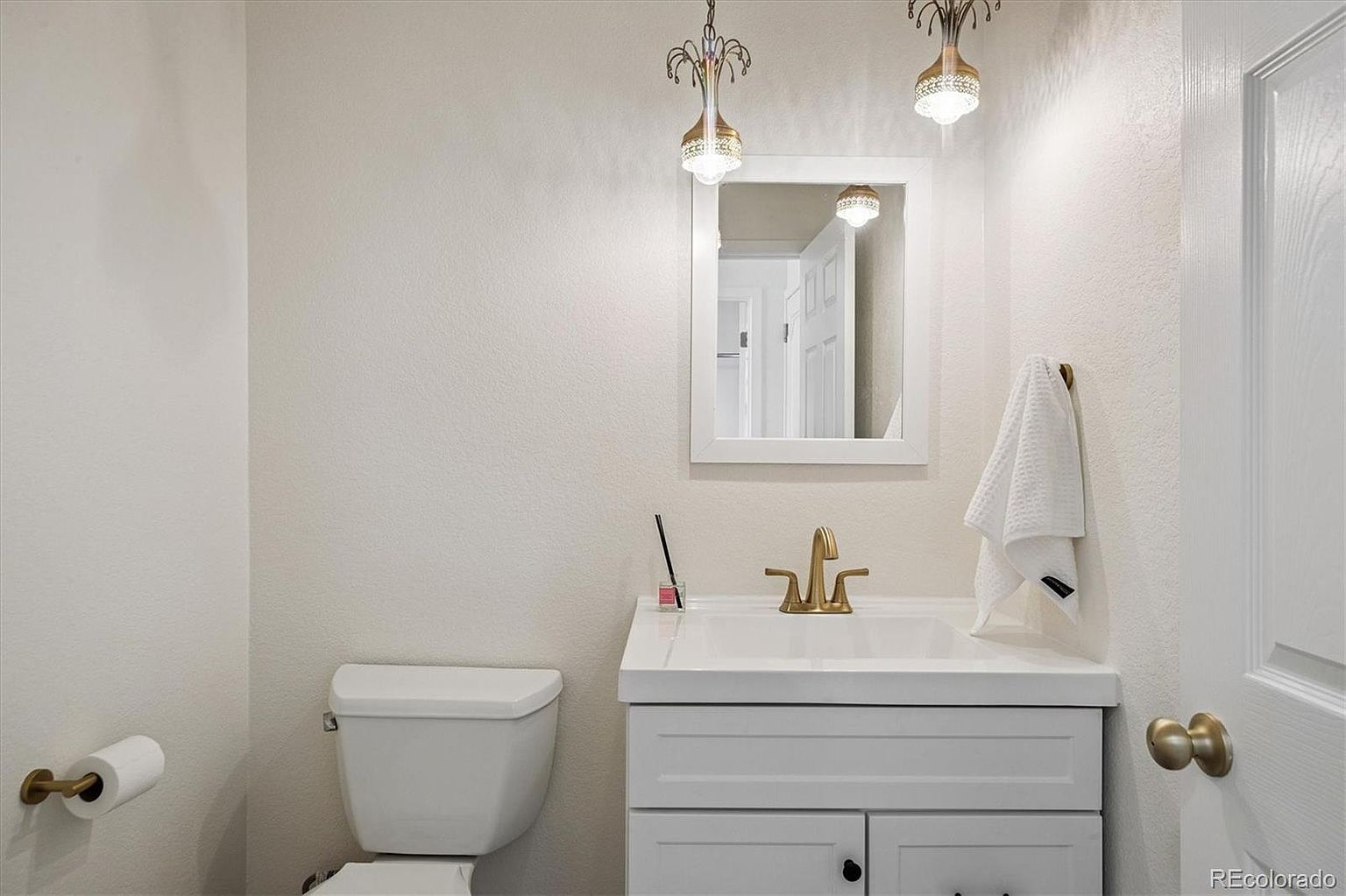 This is a well-lit guest bathroom featuring a white toilet and a vanity with a white countertop and cabinet. A gold-toned faucet and matching light fixtures add a touch of elegance. A white-framed mirror hangs above the sink, and a white towel is neatly placed on a towel rack, creating a clean and inviting space.