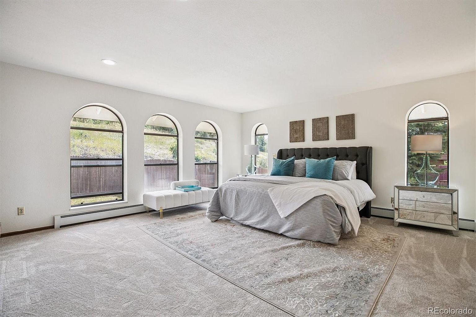 This is a spacious primary bedroom featuring a large bed with a tufted headboard, complemented by blue accent pillows and a gray comforter. The room is well-lit with natural light streaming through arched windows, and the neutral color palette creates a serene and inviting atmosphere. A decorative rug anchors the space, adding texture and visual interest.