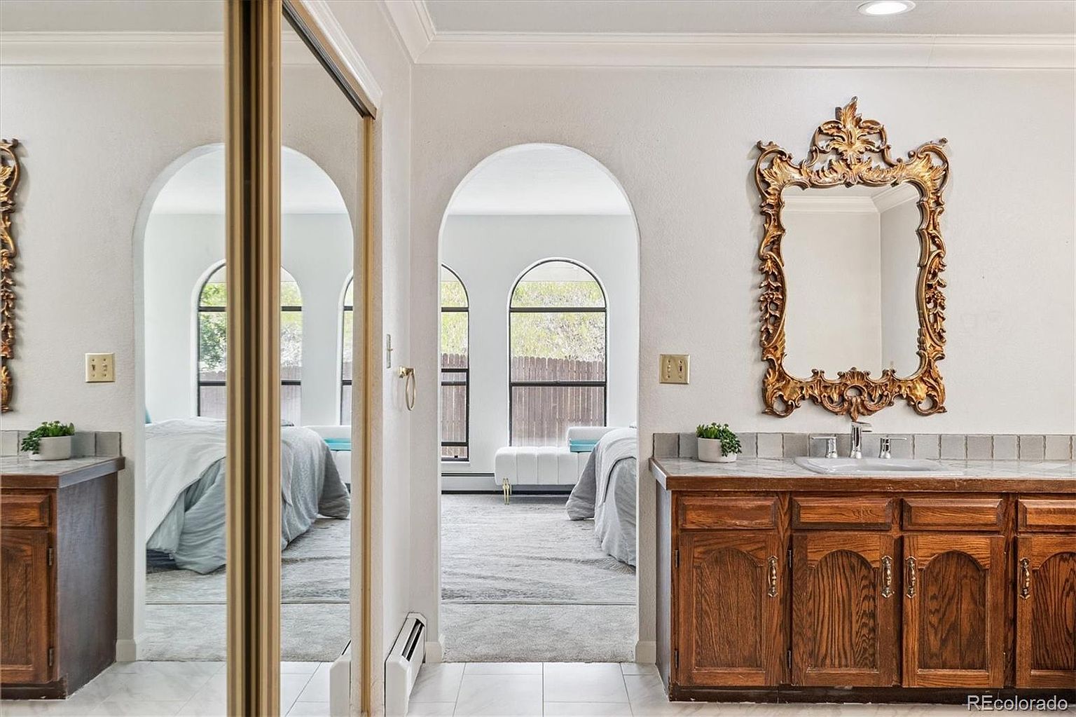 This is a primary bathroom featuring a vanity with wooden cabinets and a marble countertop, complemented by an ornate gold-framed mirror. The bathroom opens into a bedroom through an arched doorway, offering a glimpse of the bedroom's interior with its arched windows and neutral color palette. The overall style is traditional with a touch of elegance.