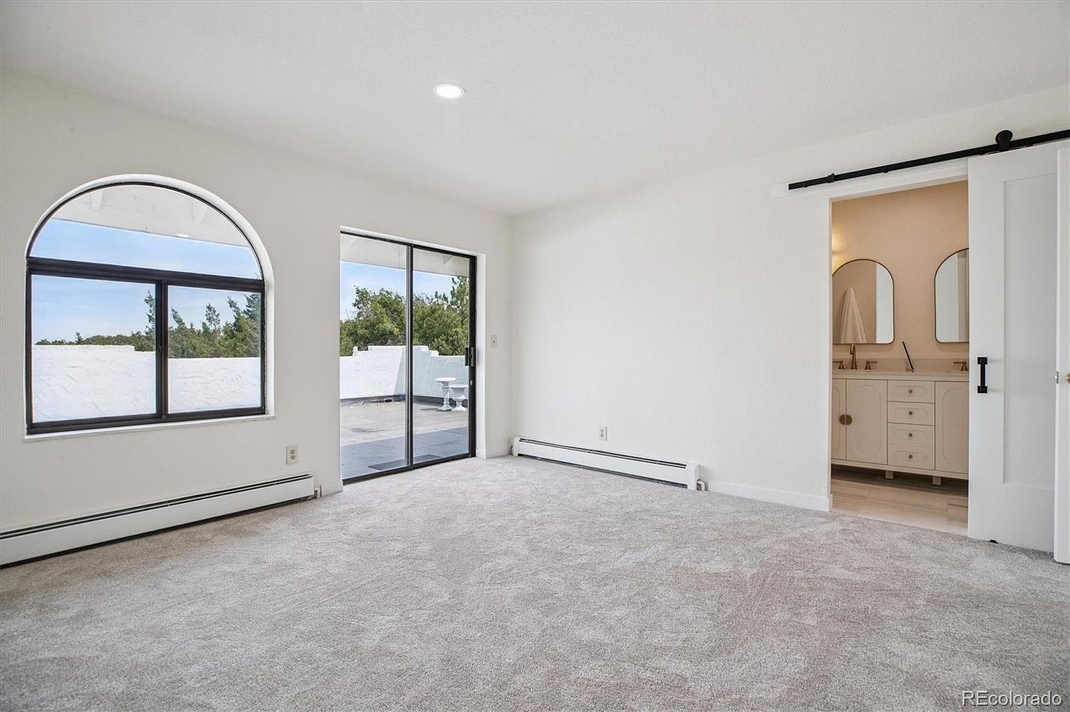 This is a bright and spacious primary bedroom featuring neutral-toned carpet, white walls, and a modern aesthetic. The room has a large arched window and sliding glass doors leading to an outdoor area, along with a sliding barn door that opens to the bathroom. The room is well-lit and appears clean and inviting.