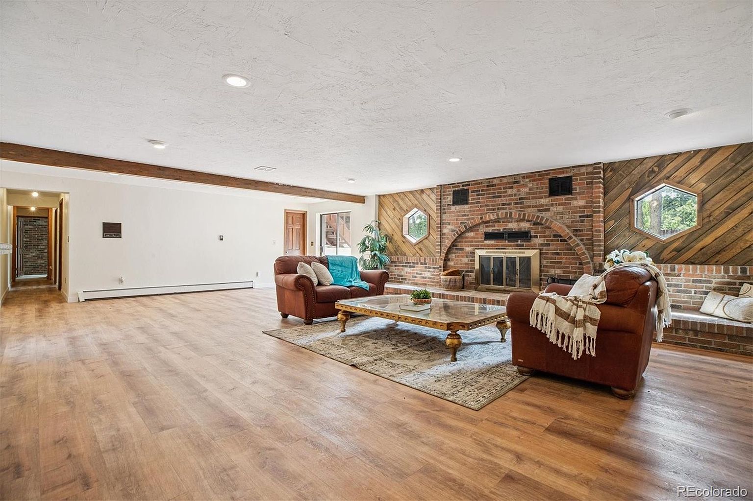 This is a spacious living room featuring hardwood floors, a brick fireplace, and comfortable leather armchairs. The room is well-lit with recessed lighting and natural light from the windows. A large coffee table sits in the center of the room, adding to the inviting atmosphere.