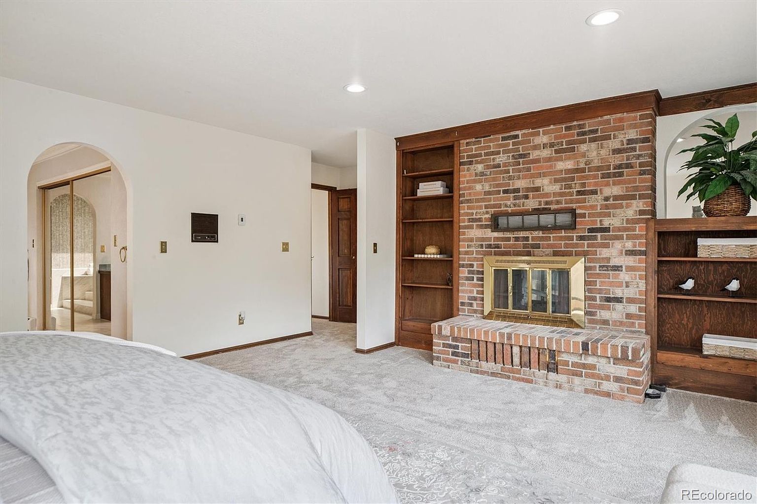 This is a primary bedroom featuring a brick fireplace with built-in bookshelves on either side. The room has neutral carpet and white walls, with a doorway leading to an en-suite bathroom. The perspective is from the foot of the bed, showcasing the room's layout and focal point.