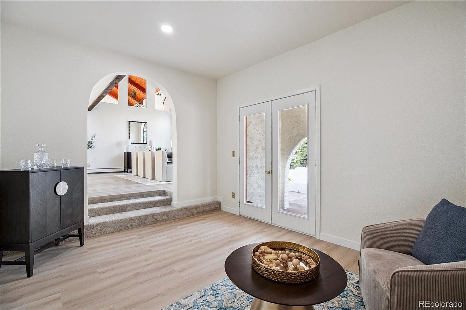 This interior shot showcases a stylish living room with a blend of modern and rustic elements. The room features light wood flooring, a dark wood cabinet, and a round coffee table with decorative elements. An arched doorway leads to another room, adding architectural interest, while double doors provide access to the outside. The overall impression is bright, inviting, and well-designed.