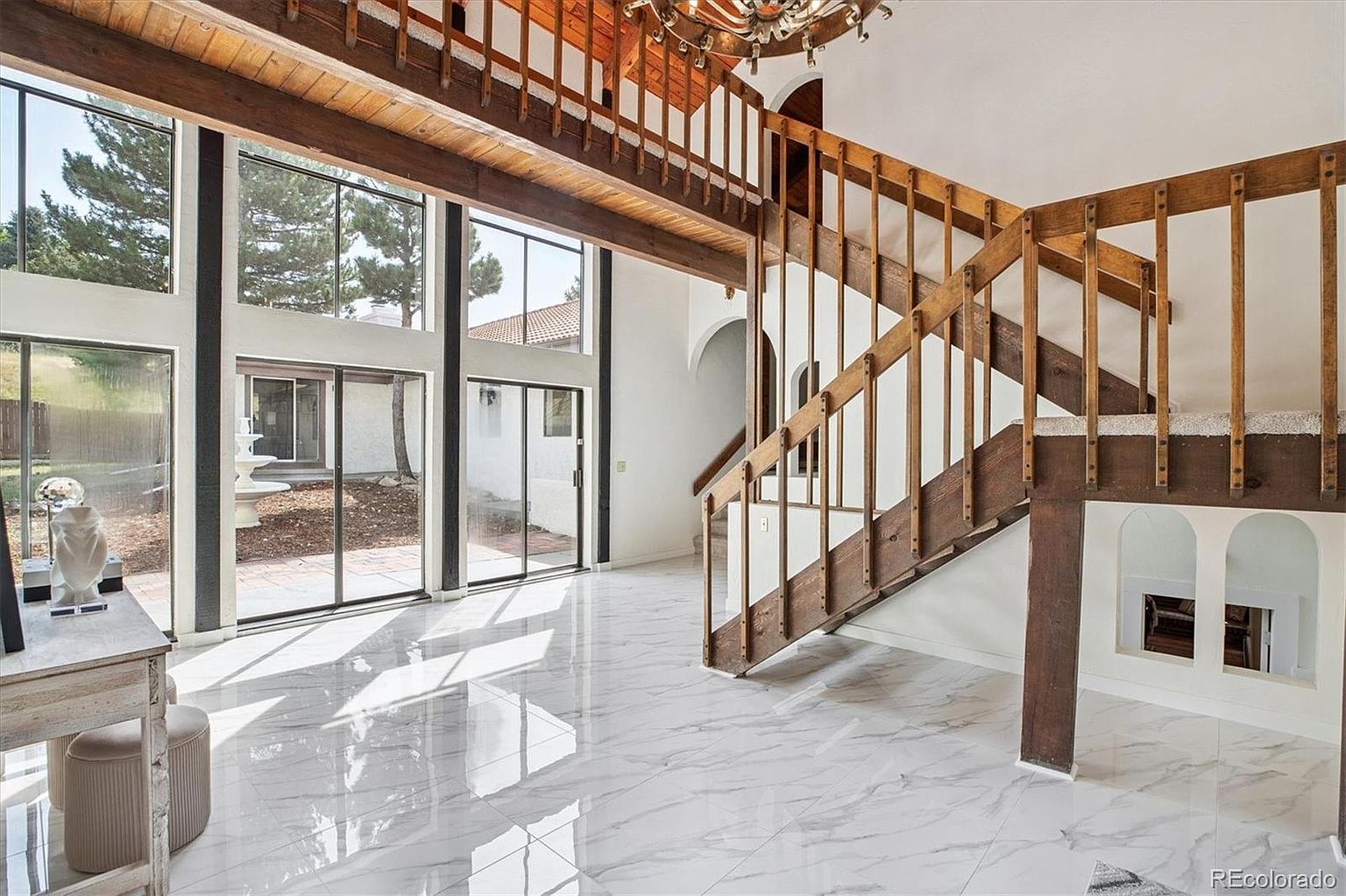 This interior shot showcases a grand hallway with a striking wooden staircase. The space is bright and airy, thanks to large windows that flood the area with natural light, reflecting off the marble flooring. The architectural design features a blend of modern and rustic elements, creating a unique and inviting atmosphere.