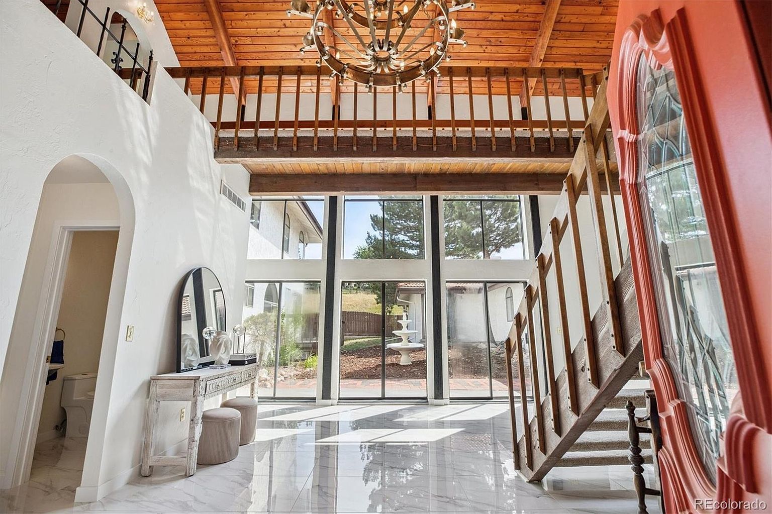 This is an interior shot of a grand foyer featuring a soaring ceiling with wooden beams and a decorative chandelier. Large windows provide ample natural light and a view of the exterior. A staircase with wooden railings is visible on the right, and a hallway leads to other rooms on the left. The flooring is polished marble, adding to the luxurious feel of the space.