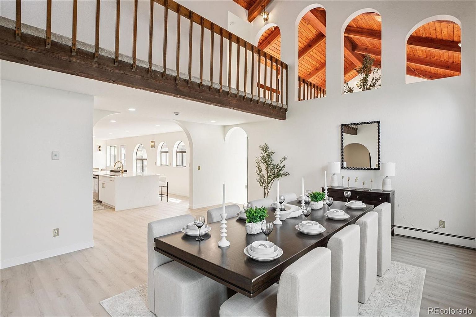 This interior shot showcases a dining room with a large, dark wood table set for a meal, surrounded by light gray upholstered chairs. The room features high ceilings with arched windows and a balcony overlooking the space, creating an open and airy atmosphere. A dark wood sideboard with a mirror above it adds a touch of elegance to the room.