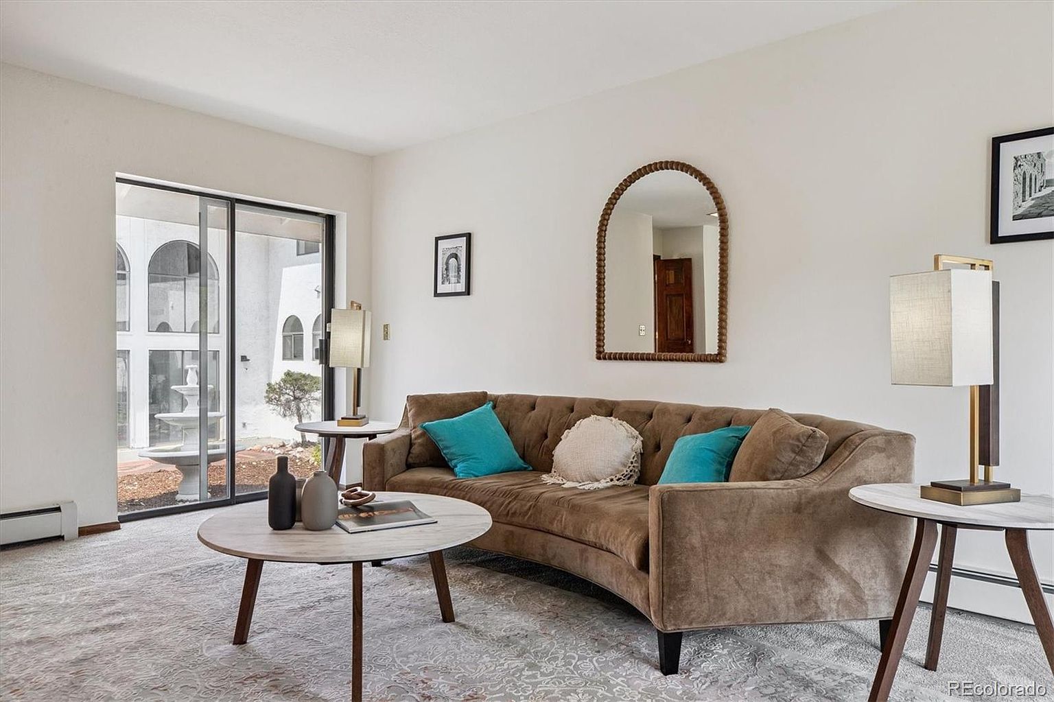 This is an interior shot of a living room featuring a curved brown sofa with blue pillows, a round coffee table with decorative items, and two side tables with lamps. A large sliding glass door provides natural light and a view of an outdoor fountain, while a decorative mirror and framed artwork adorn the walls. The room has a cozy and inviting atmosphere.