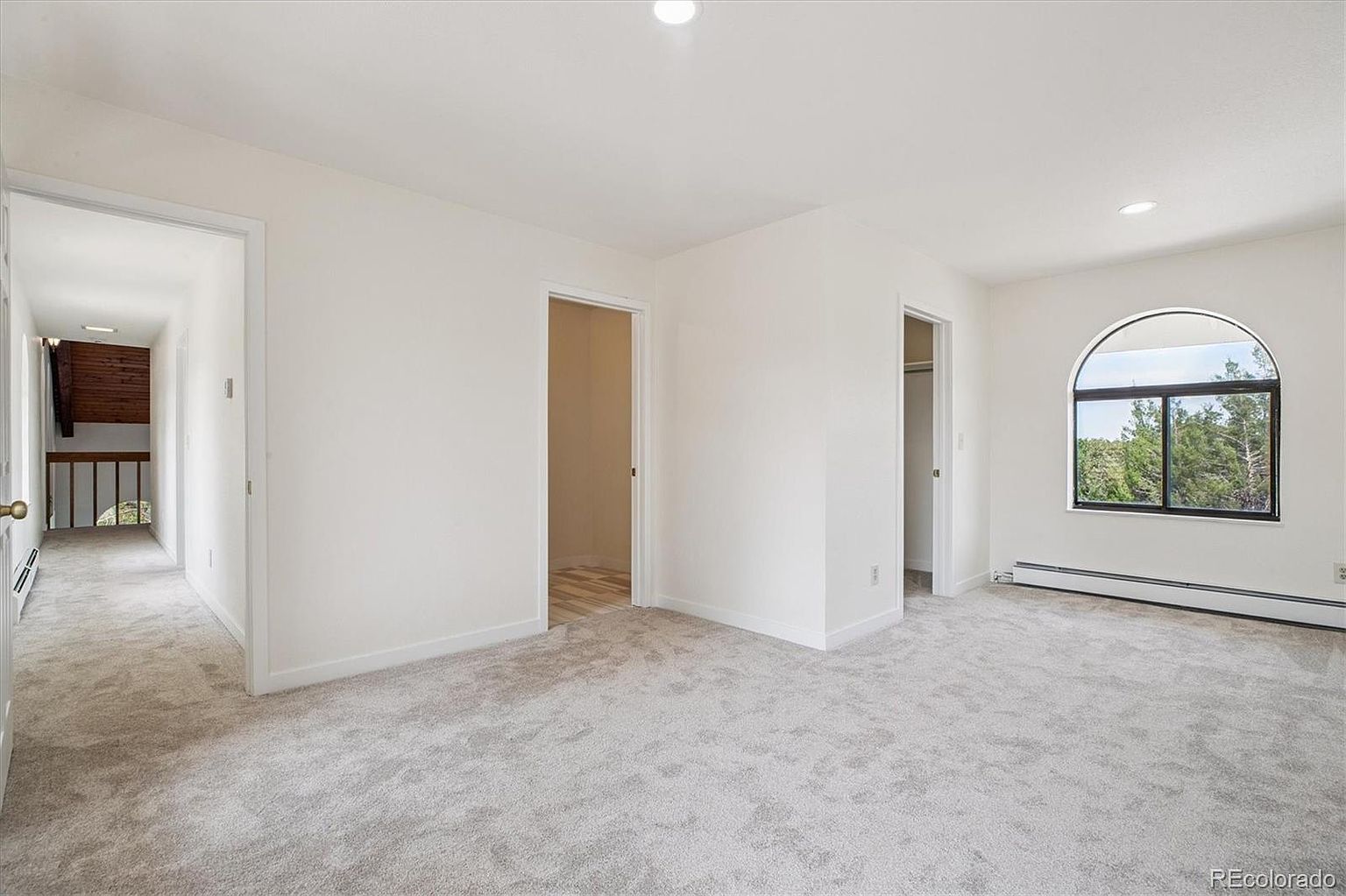 This is a bright and spacious primary bedroom featuring neutral carpeting and white walls. The room has an arched window providing natural light and views of the outdoors. There are also two doorways leading to other areas, possibly a hallway and a closet or bathroom, enhancing the flow of the space.