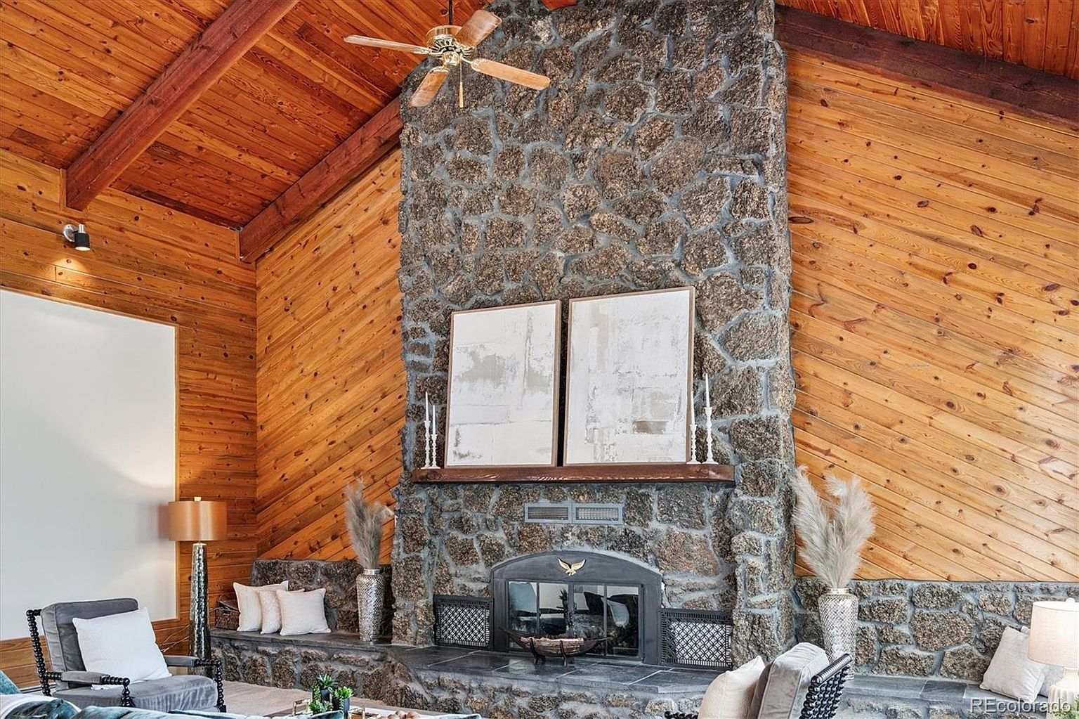 This interior shot showcases a living room with a prominent stone fireplace, flanked by wood-paneled walls and ceiling. The room features a rustic yet elegant style, with neutral-toned furniture and decor. The fireplace serves as a focal point, adding warmth and character to the space.