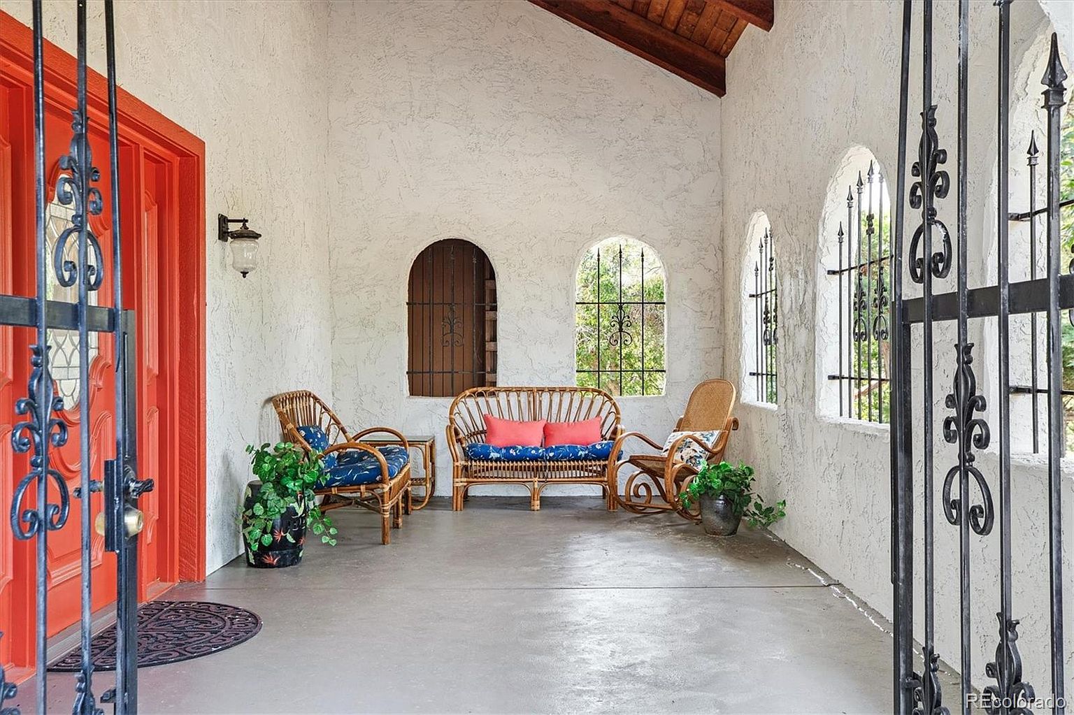 This image showcases a charming outdoor patio area, featuring a vibrant orange front door and decorative wrought iron gate. The patio is furnished with wicker seating, including a loveseat and chairs, adorned with patterned cushions. Arched windows with wrought iron details provide natural light and a view of the surrounding greenery, creating a cozy and inviting space.