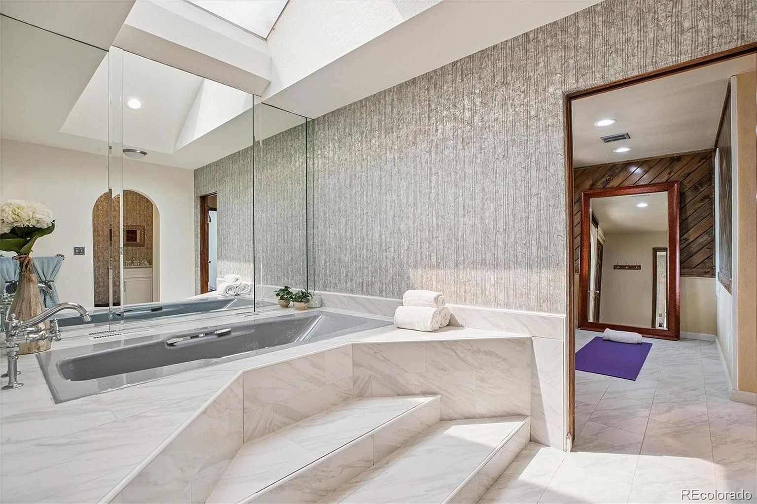This is a luxurious primary bathroom featuring a large, gray-toned bathtub with marble steps leading up to it. The walls are adorned with textured wallpaper, and a large mirror reflects the space, creating an open and airy feel. A doorway leads to another room, showcasing wood paneling and a full-length mirror, adding depth and interest to the scene.