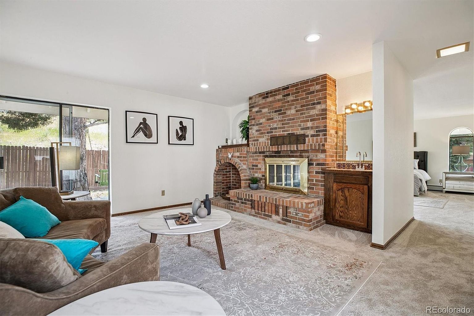This is an interior shot of a living room featuring a prominent brick fireplace. The room is furnished with comfortable brown sofas accented by teal pillows, and a round coffee table sits on a patterned rug. The space is well-lit and appears inviting, with a glimpse into an adjacent room suggesting an open floor plan.