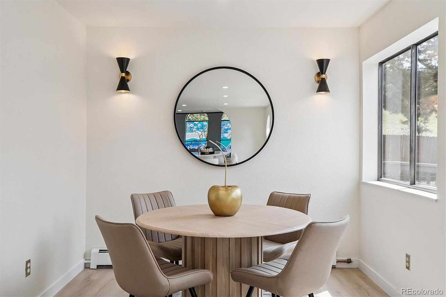This dining room features a round wooden table with a unique pedestal base, surrounded by four upholstered chairs. A round mirror hangs on the wall, flanked by two sconce lights, adding a touch of modern elegance. Natural light streams in from a nearby window, creating a bright and inviting atmosphere.