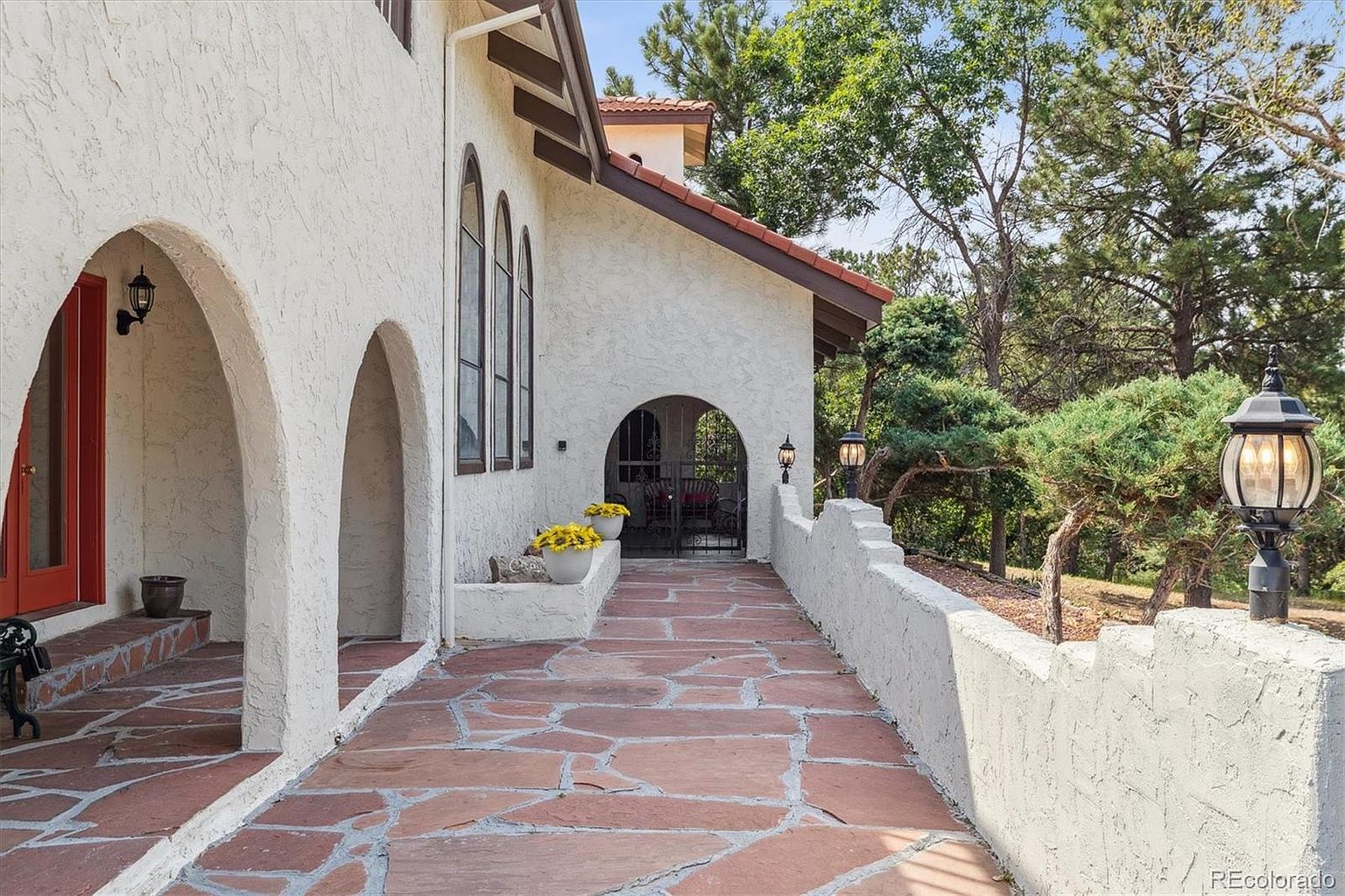 This image showcases the entryway of a home, featuring a charming walkway paved with red flagstone leading to an arched gate. The exterior walls are textured white stucco, complemented by arched architectural details and dark-framed windows. The scene is framed by lush greenery and decorative outdoor lighting, creating an inviting and elegant first impression.
