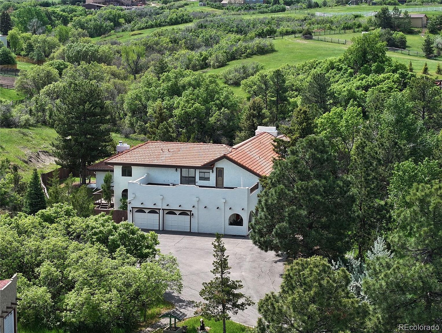 This aerial view showcases a beautiful white stucco home with a red tile roof, nestled among lush greenery and mature trees. The property features a spacious driveway leading to a two-car garage, and the surrounding landscape includes rolling hills and a well-maintained lawn. The overall impression is one of privacy and tranquility in a desirable location.
