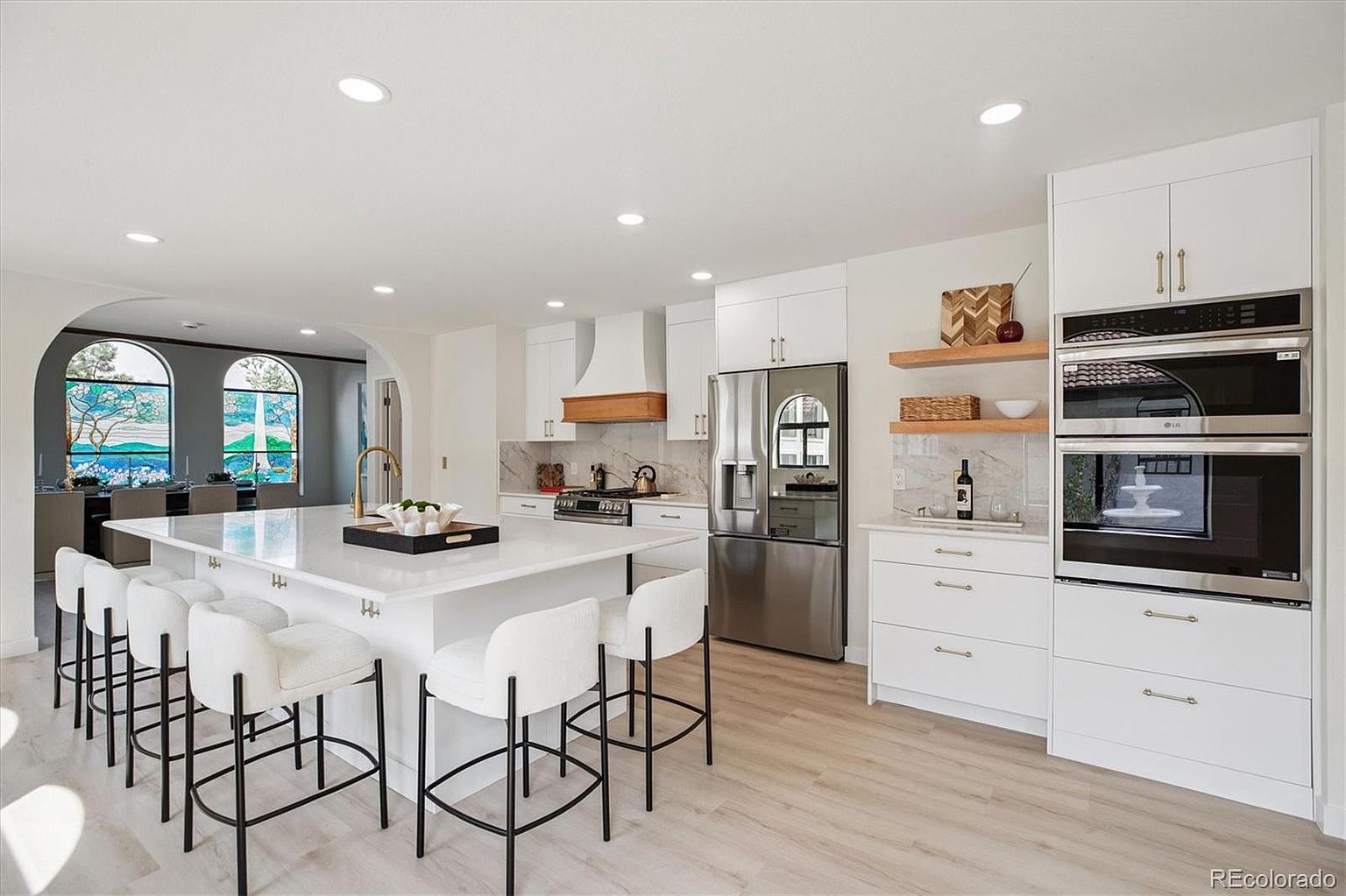 This is a bright and modern kitchen featuring white cabinetry, stainless steel appliances, and a large central island with seating. The kitchen is open to a dining area with arched windows, creating a spacious and inviting atmosphere. The design incorporates clean lines and a neutral color palette, enhancing the sense of openness and sophistication.
