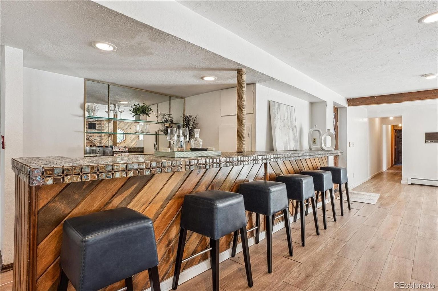 This interior shot showcases a stylish basement bar area. The bar features a unique wood design with a tiled countertop, complemented by several black leather bar stools. A mirrored backdrop with glass shelving adds depth and sophistication, while the wood flooring and neutral walls create a warm and inviting atmosphere.