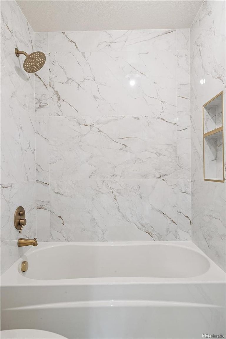 This is a well-lit bathroom featuring a white bathtub with bronze fixtures. The walls are covered in large, marble-patterned tiles, and a built-in shelf provides storage. The overall impression is clean and modern, with a touch of elegance.