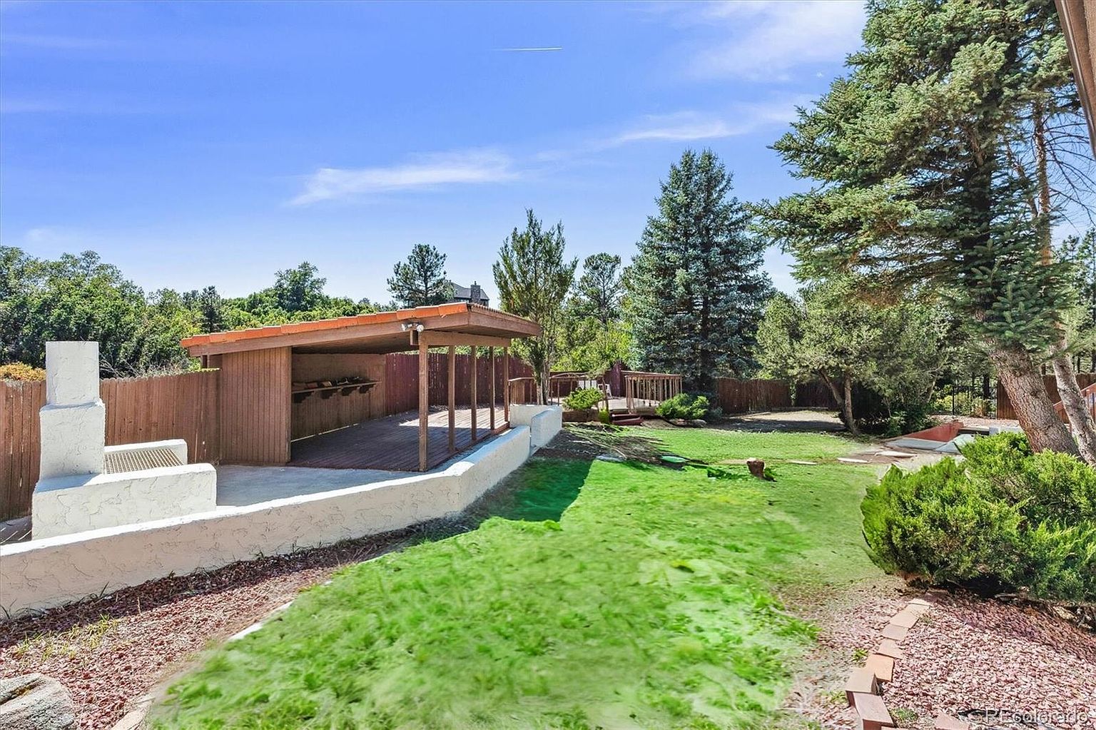 This image showcases a well-maintained backyard featuring a covered patio area with a built-in grill, a lush green lawn, and mature trees providing shade and privacy. A white retaining wall adds architectural interest, and a wooden deck extends the outdoor living space. The overall impression is one of a serene and inviting outdoor retreat.