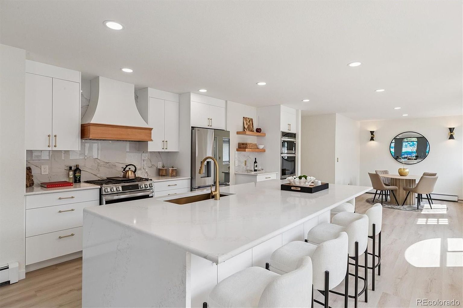 This is a bright and modern kitchen featuring white cabinetry, stainless steel appliances, and a large island with a white countertop and seating. The kitchen also includes a dining area with a round table and chairs, creating an open and inviting space. The perspective is wide, showcasing the kitchen's layout and connection to the dining area.