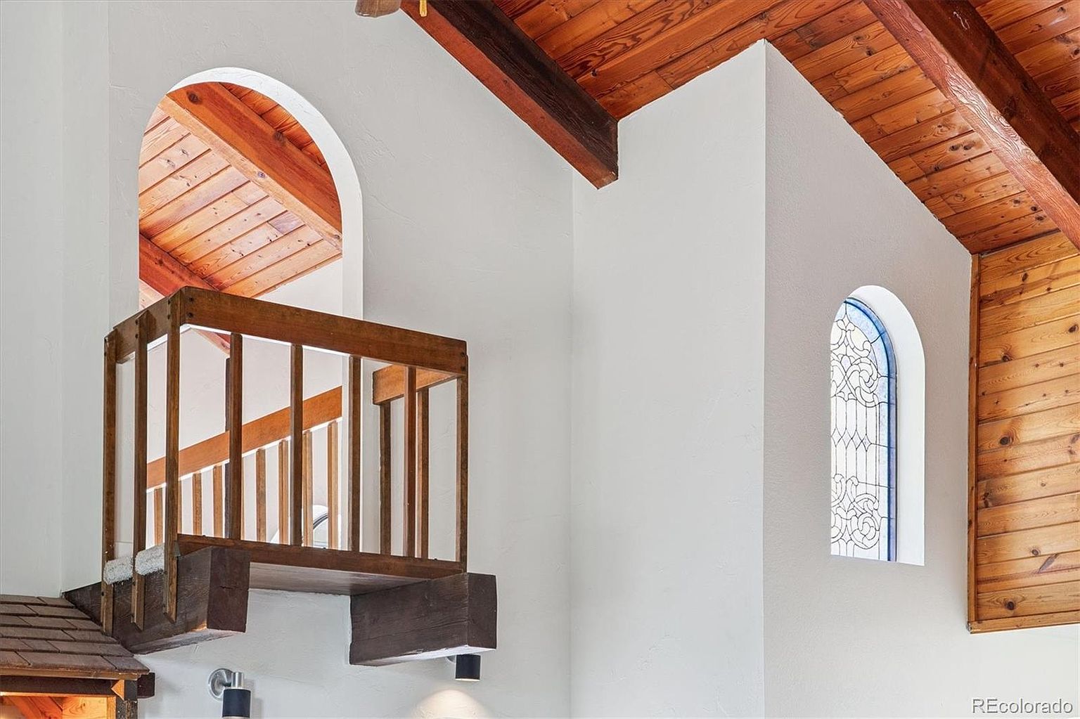 This image showcases a unique architectural detail within a home, featuring a small interior balcony with wooden railings and support beams. An arched window and a stained-glass window add character to the white walls, while the exposed wooden ceiling beams contribute to a rustic yet elegant aesthetic. The perspective is from below, looking up at the balcony and windows.