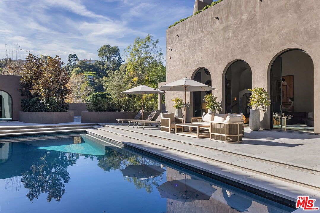 This serene outdoor living space features a large, rectangular swimming pool set against a Mediterranean-style home with arched doorways and a textured, neutral-toned facade. The stone-paved patio is furnished with modern lounge chairs and a comfortable seating area shaded by umbrellas, creating an inviting atmosphere for relaxation. The scene is framed by lush greenery and trees, offering a private and tranquil backyard retreat.