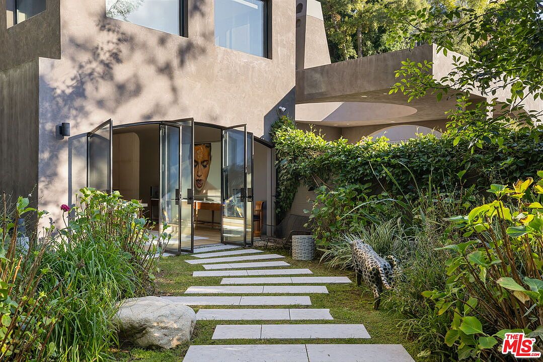 This serene garden scene features a modern home exterior with large glass doors opening onto a stone-paved walkway. The lush, landscaped yard includes a decorative leopard sculpture, dense greenery, and a unique architectural overhang that creates a shaded transition space. The perspective is at eye level, capturing a tranquil and sophisticated outdoor living area.
