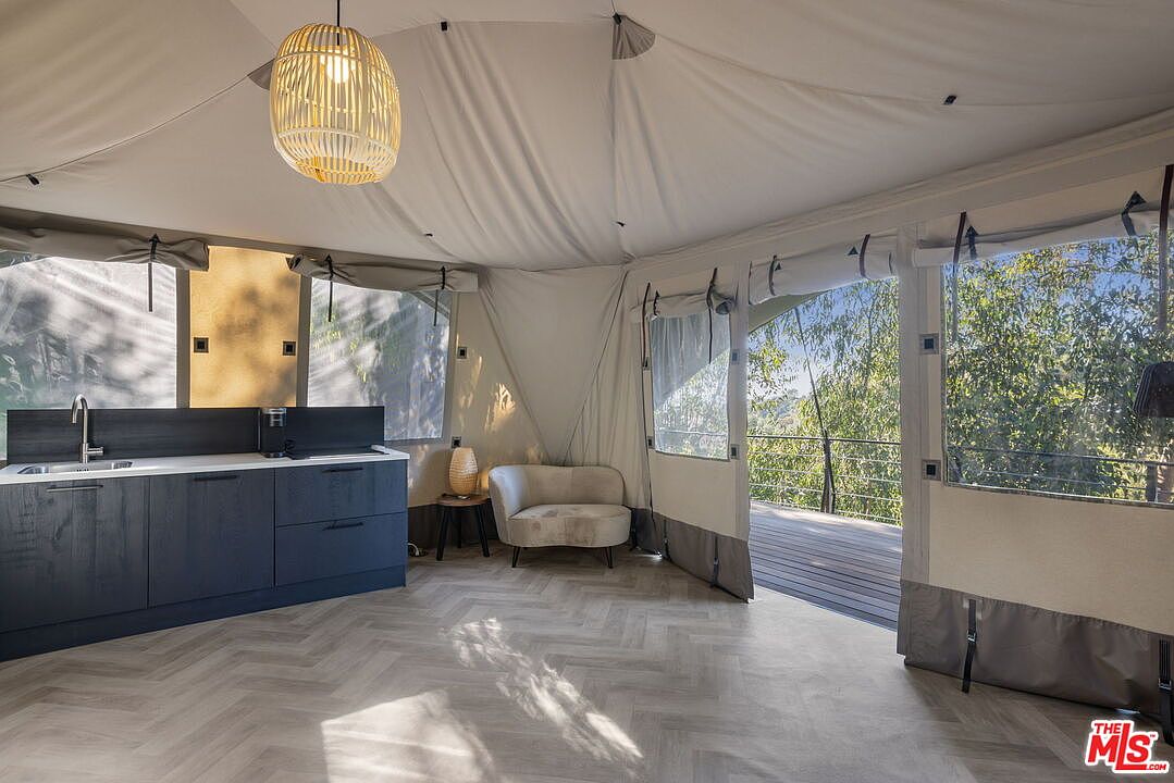 This unique interior space features a tent-like structure with soft, draped fabric walls and a light-toned herringbone floor. A modern dark-toned kitchenette sits along one wall, while a plush, neutral-colored armchair is tucked into the corner near a large opening that leads to an outdoor deck. The room is illuminated by a woven pendant light, creating a serene and airy atmosphere that blends indoor comfort with an outdoor aesthetic.