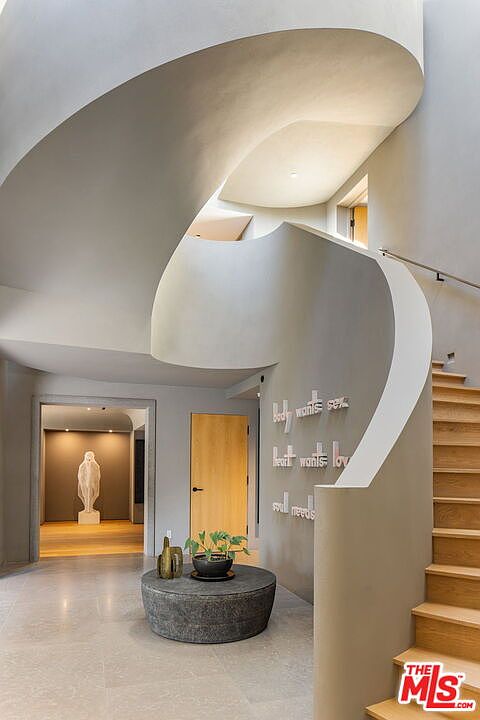 This striking entryway features a dramatic, sculptural curved staircase with a smooth, light-toned finish that draws the eye upward. A circular stone table sits in the foreground, topped with a small potted plant, while a doorway in the background reveals a gallery-like space with a tall, abstract sculpture. The wall beside the stairs is adorned with three-dimensional lettering, adding a unique artistic touch to the minimalist, modern architectural design.