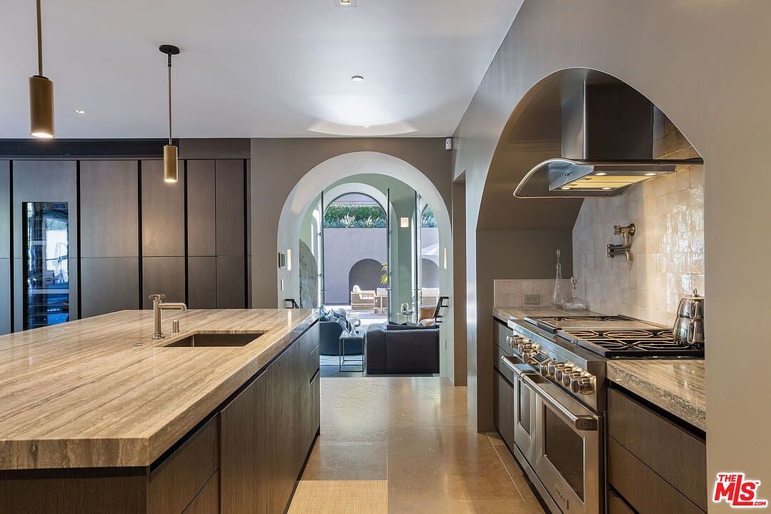 This sophisticated kitchen features a large central island with a prominent stone countertop and a built-in sink, complemented by dark cabinetry and high-end stainless steel appliances. An arched opening provides a clear sightline through to an adjacent living area and an outdoor patio space, creating a seamless transition between indoor and outdoor living. The design aesthetic is modern and refined, utilizing neutral tones and clean lines to create an inviting, open-concept atmosphere.