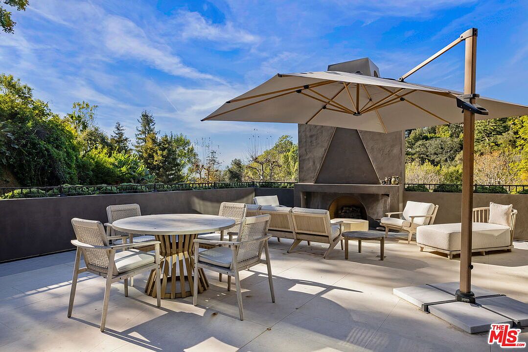 This spacious outdoor terrace features a modern dining set and a comfortable lounge area arranged around a tall, minimalist outdoor fireplace. A large cantilevered umbrella provides shade over the dining table, while the open-air setting offers a clear view of the surrounding lush greenery and blue sky. The neutral-toned stone flooring and contemporary furniture create a sophisticated and inviting space for al fresco entertaining.
