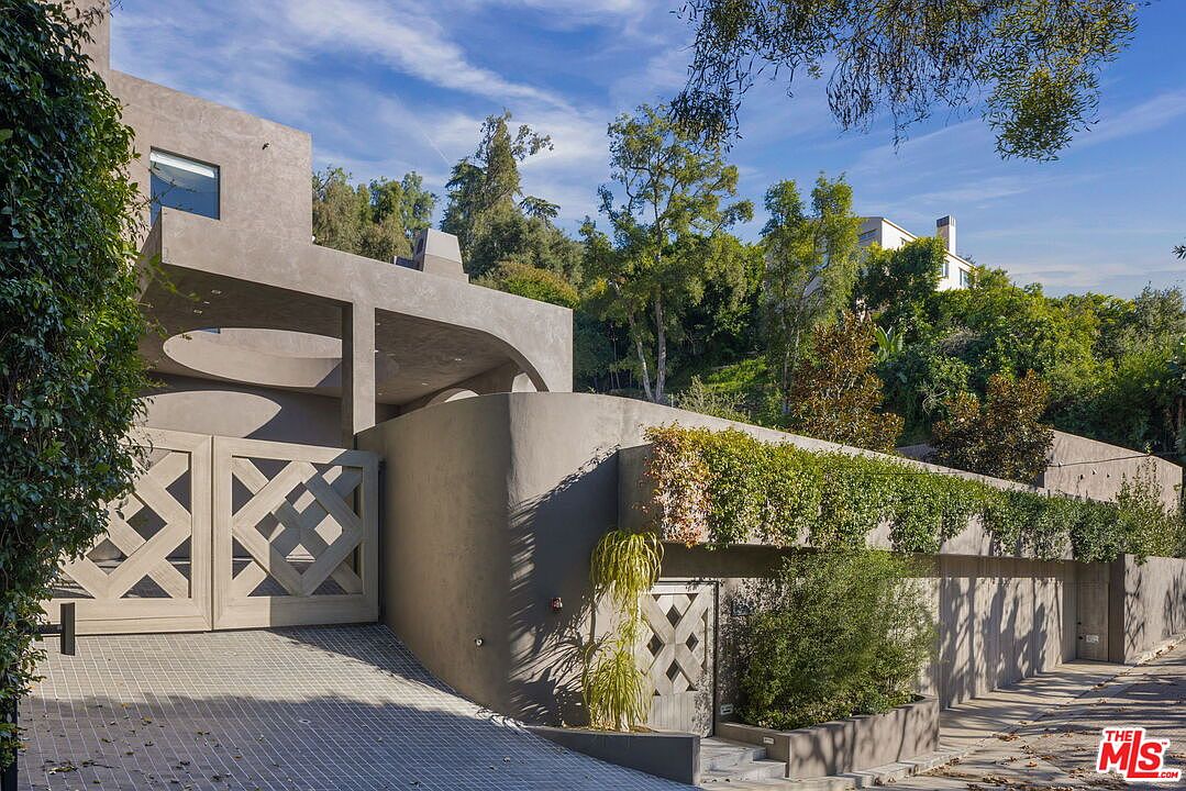 This contemporary residence features a striking, minimalist architectural design with smooth, grey stucco walls and geometric, lattice-patterned gates. The property is nestled into a hillside, accented by lush greenery and a long, horizontal planter box overflowing with vines along the upper wall. The perspective captures the gated driveway and the unique, sculptural curves of the building's facade under a clear blue sky.