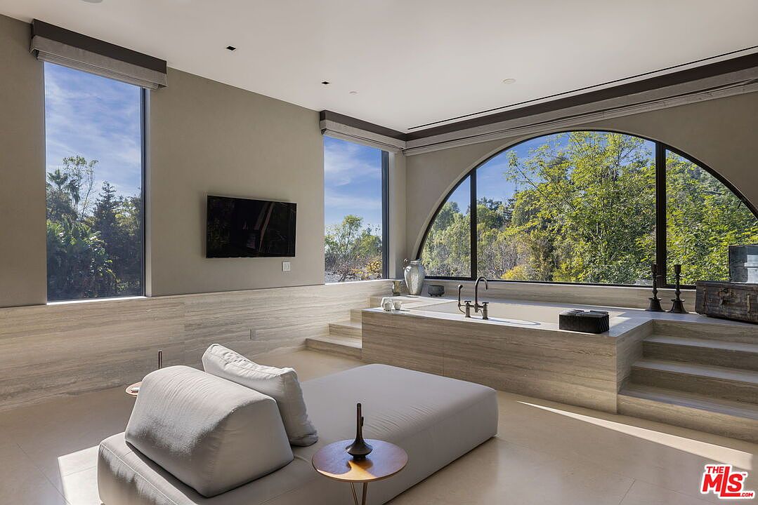 This luxurious primary bathroom features a sunken soaking tub set within a raised stone platform, positioned beneath a large arched window that offers expansive views of the surrounding trees. A minimalist lounge chair sits in the foreground, creating a serene, spa-like atmosphere. The room is finished with neutral stone walls and flooring, complemented by recessed lighting and a wall-mounted television.