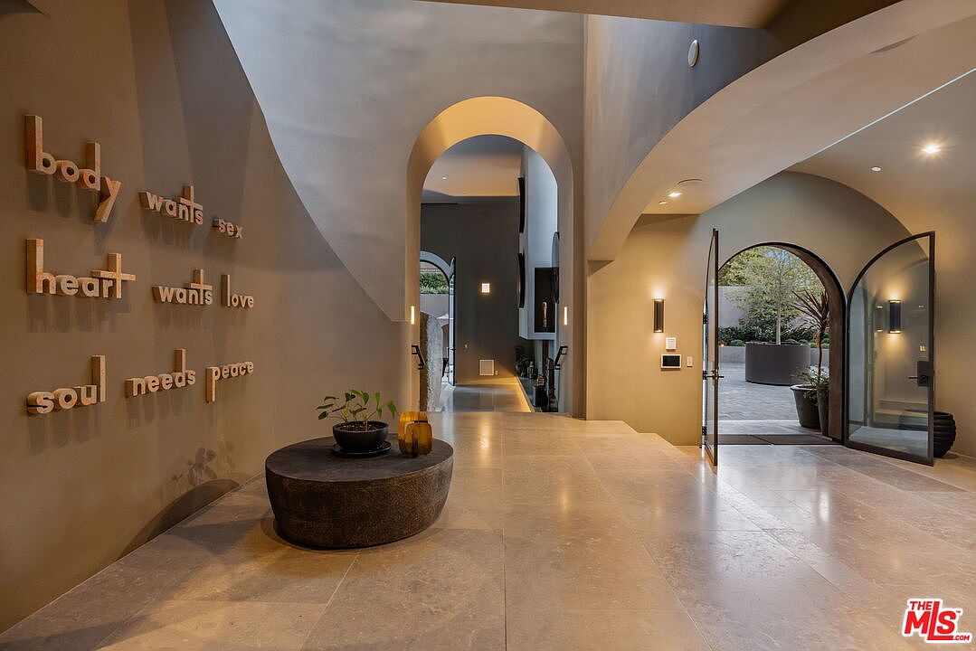 This elegant entryway features a spacious, open-concept design with smooth, neutral-toned walls and large stone floor tiles. A prominent wall display showcases three-dimensional lettering that reads 'body wants sex, heart wants love, soul needs peace' above a minimalist stone pedestal table. The perspective looks through a grand arched transition into a secondary hallway, while an open arched doorway on the right provides a seamless connection to an outdoor courtyard.