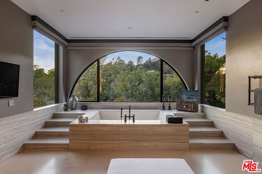 This luxurious primary bathroom features a central sunken bathtub encased in light-toned stone, accessible via steps on either side. A large, arched window serves as the focal point, framing a lush, verdant outdoor view and flooding the space with natural light. The room is finished with neutral, textured wall coverings and minimalist fixtures, creating a serene and sophisticated spa-like atmosphere.