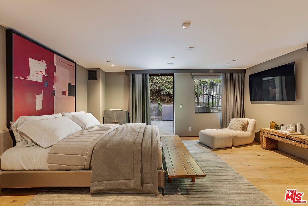 This spacious primary bedroom features a large bed with neutral bedding positioned against a wall adorned with a bold, abstract red and white art piece. The room includes a wooden bench at the foot of the bed, a comfortable lounge chair with an ottoman, and a rustic wooden console table. Large sliding glass doors open to an outdoor patio, allowing natural light to fill the room, which is finished with light wood flooring and a textured area rug.
