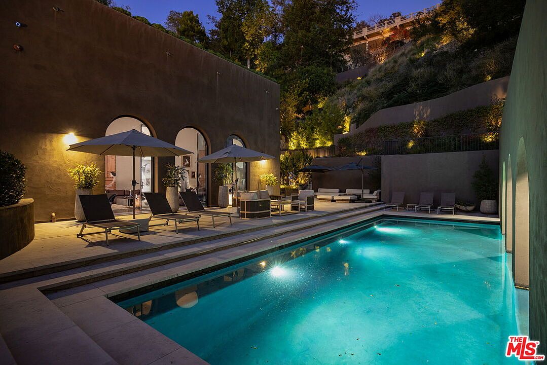 This evening shot captures a luxurious backyard pool area featuring a rectangular swimming pool illuminated by underwater lighting. The surrounding stone patio is furnished with lounge chairs and umbrellas, set against a backdrop of a multi-level hillside landscape with integrated accent lighting. The architecture features arched doorways leading into the home, creating a sophisticated and tranquil outdoor living space.