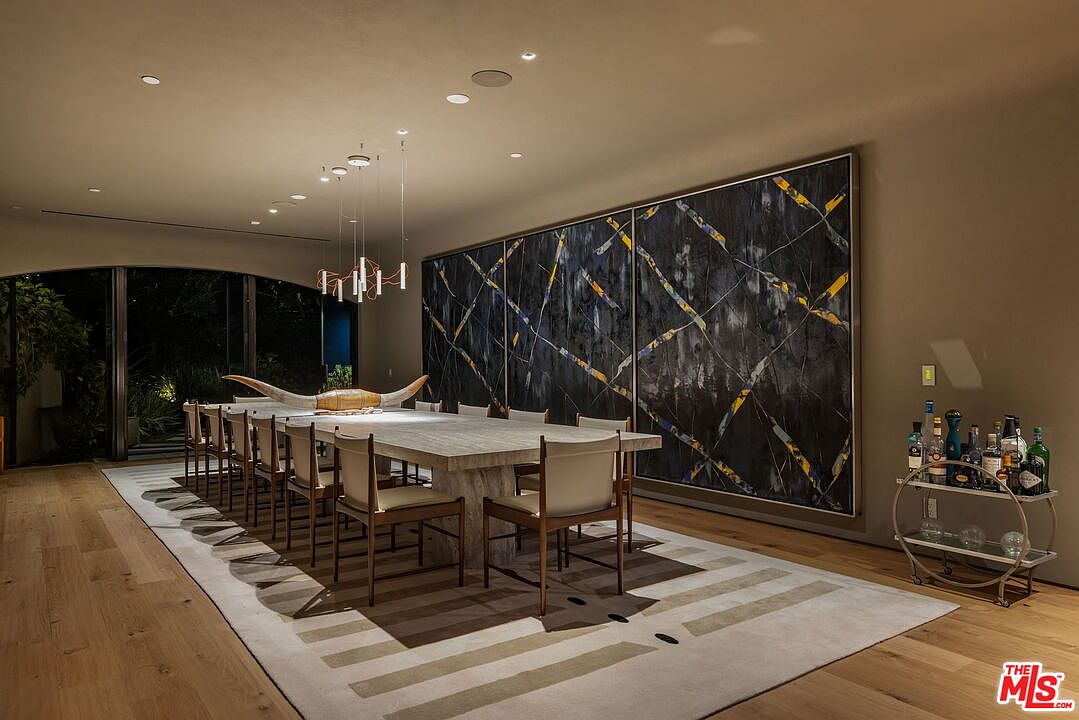 This sophisticated dining room features a long, light-toned stone table surrounded by modern chairs, set atop a large, patterned area rug. A striking, oversized abstract triptych hangs on the wall, complemented by a minimalist bar cart and a unique horn-shaped centerpiece. The space is illuminated by a series of sleek, thin pendant lights and recessed lighting, creating an elegant and contemporary atmosphere.